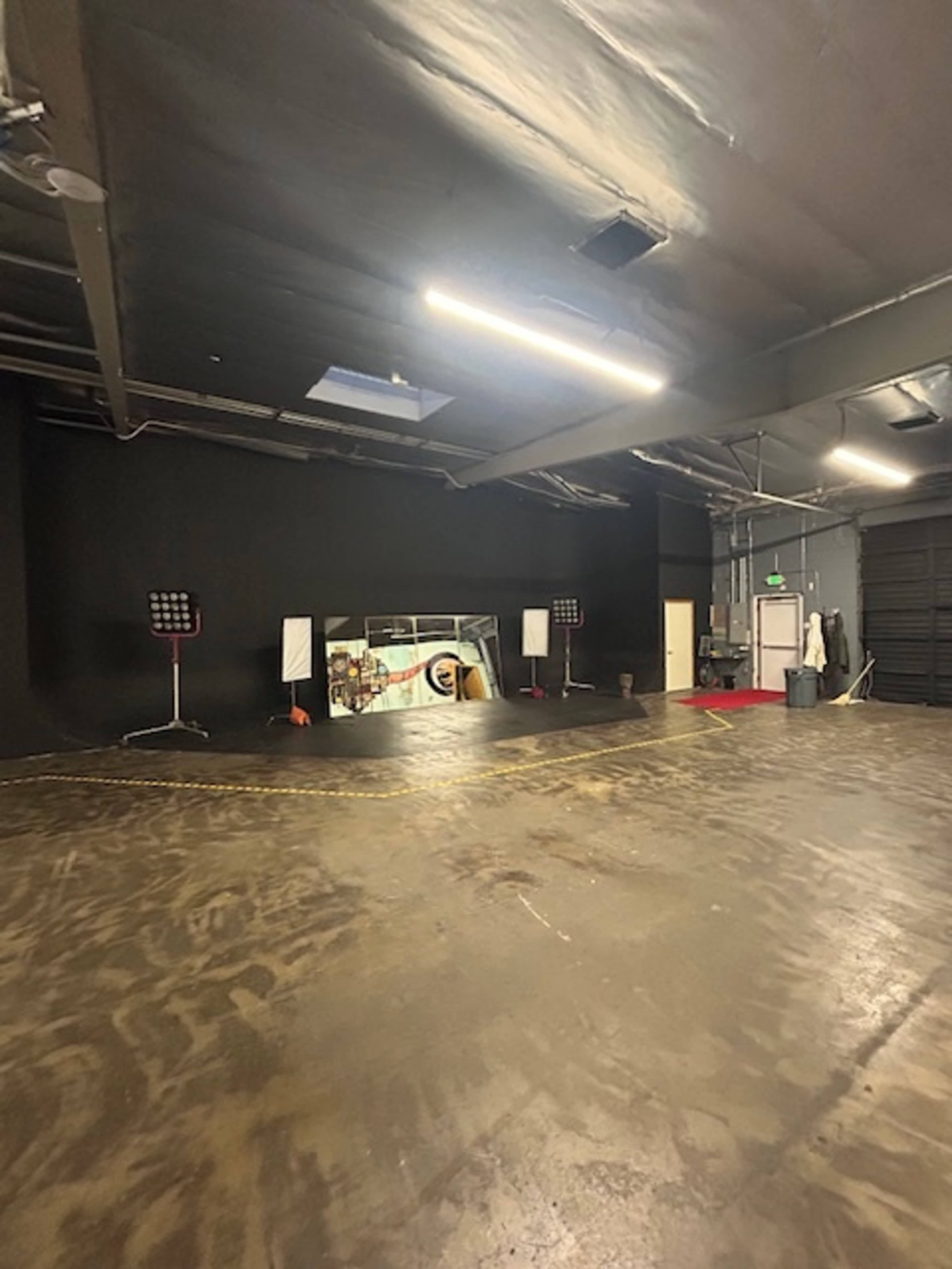 The image shows a spacious black-walled industrial studio with a polished concrete floor, featuring stage lights and a large mirror at the far end.
