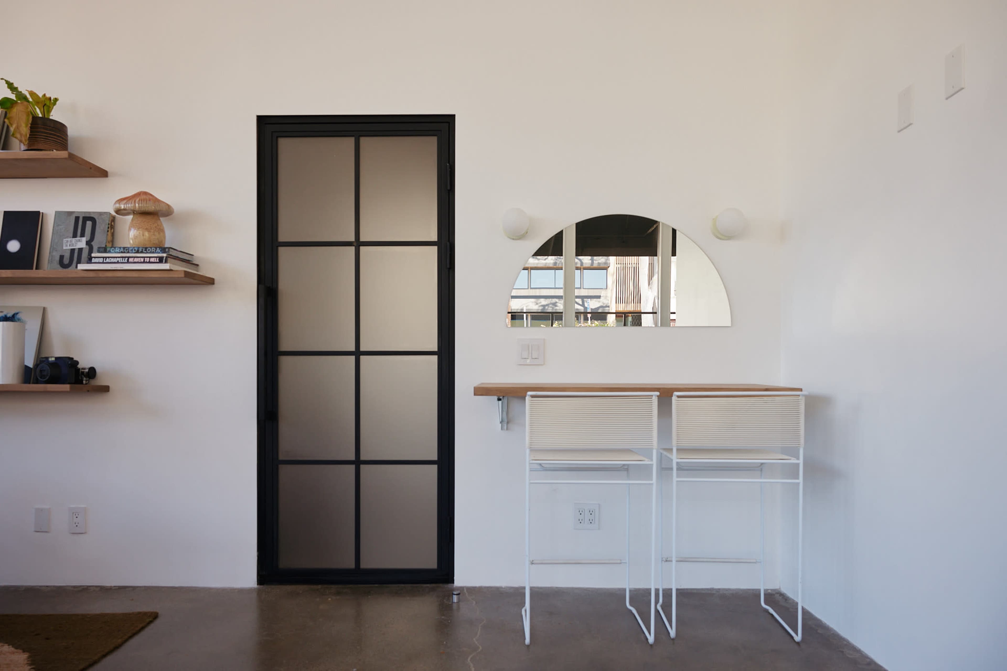 The image shows a modern interior with a minimalist design, featuring a black framed door, a wall-mounted mirror, and two white bar stools at a light wooden countertop.