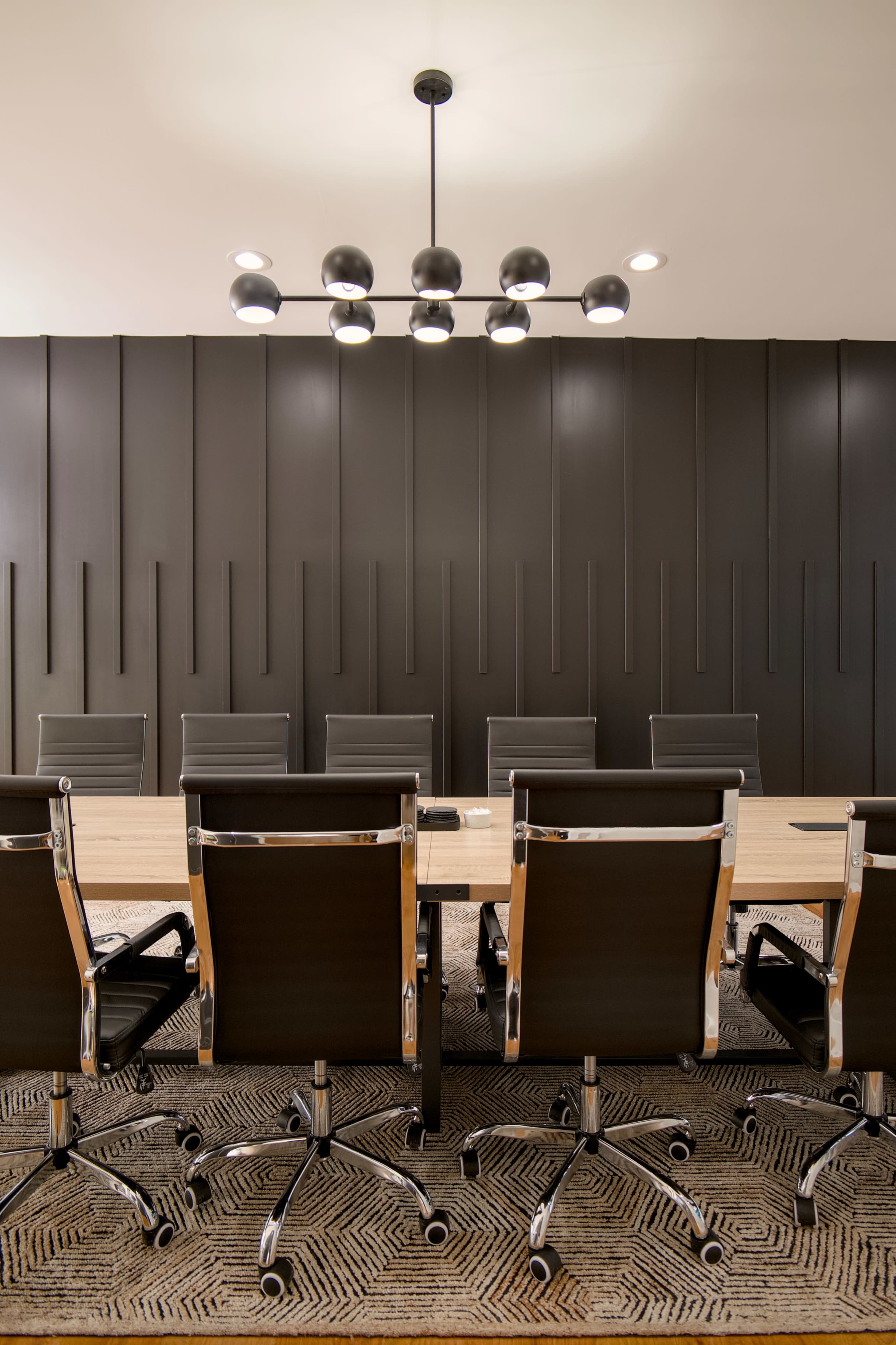 A modern conference room features a long wooden table surrounded by black swivel chairs, with a geometric chandelier overhead and a textured dark wall.