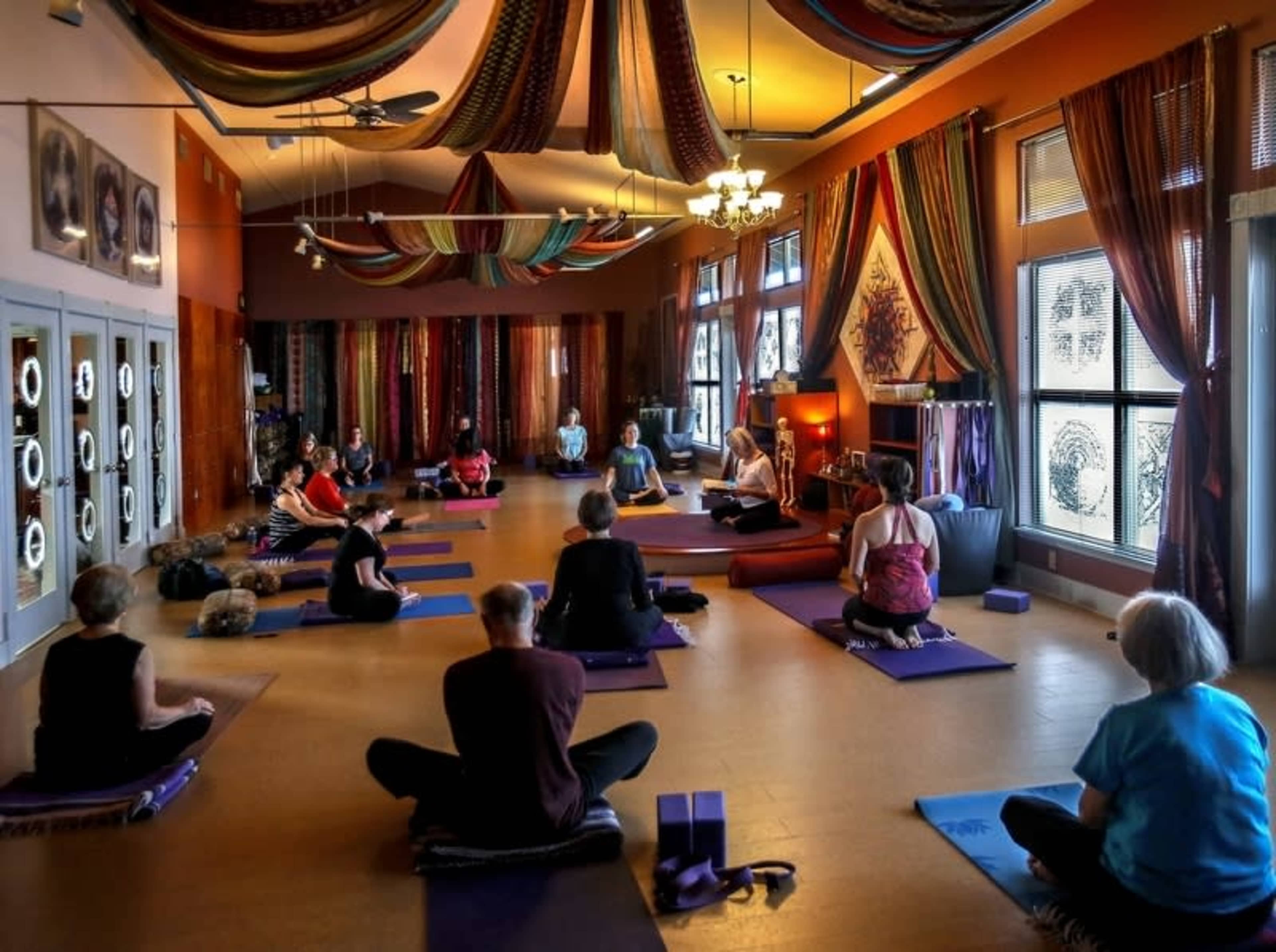 A group of people participates in a yoga class in a warmly decorated studio, seated on mats and facing an instructor at the front.