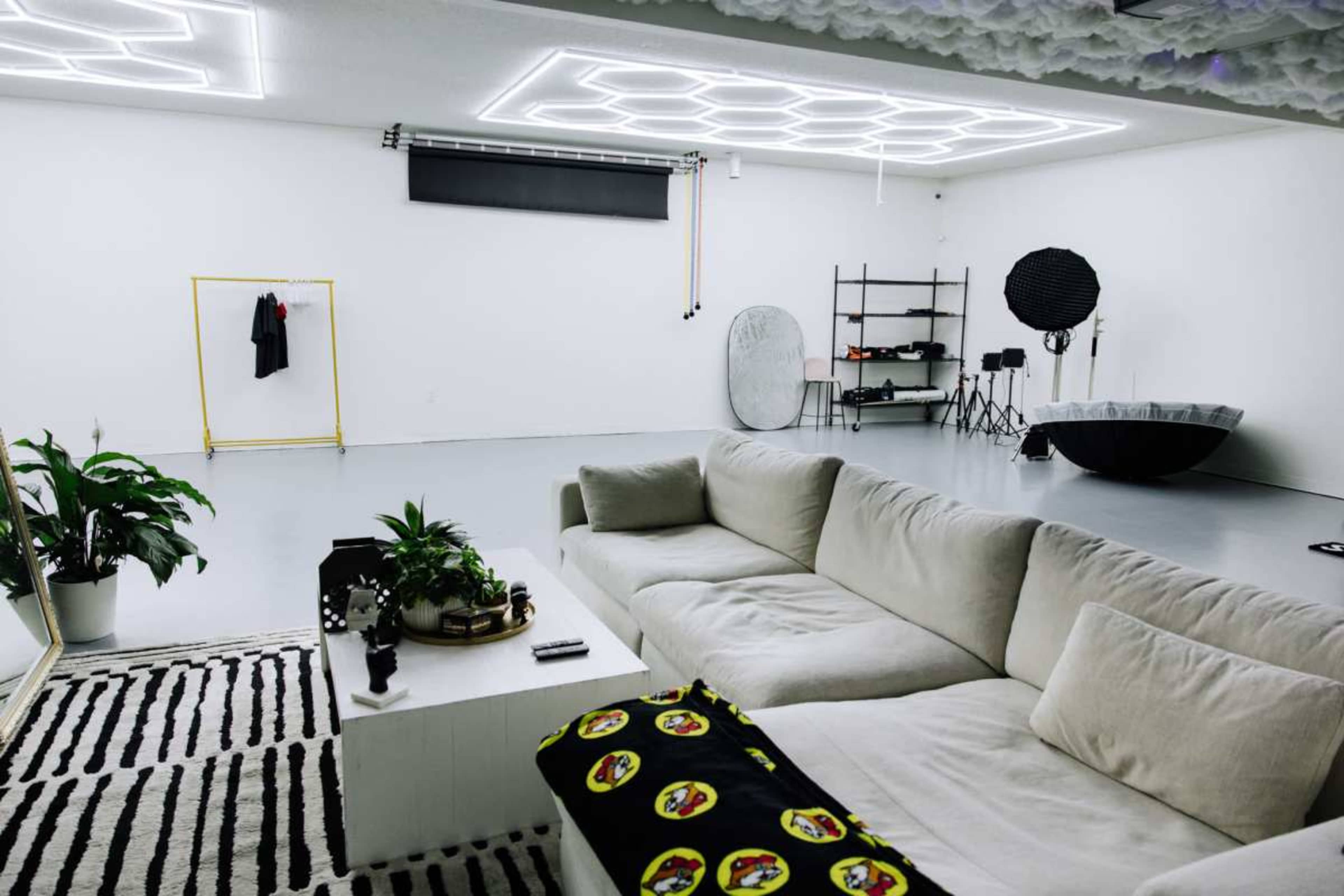 The image shows a spacious, minimalist studio featuring a white sofa, a plant, and a set of photography equipment against white walls with geometric ceiling lights.