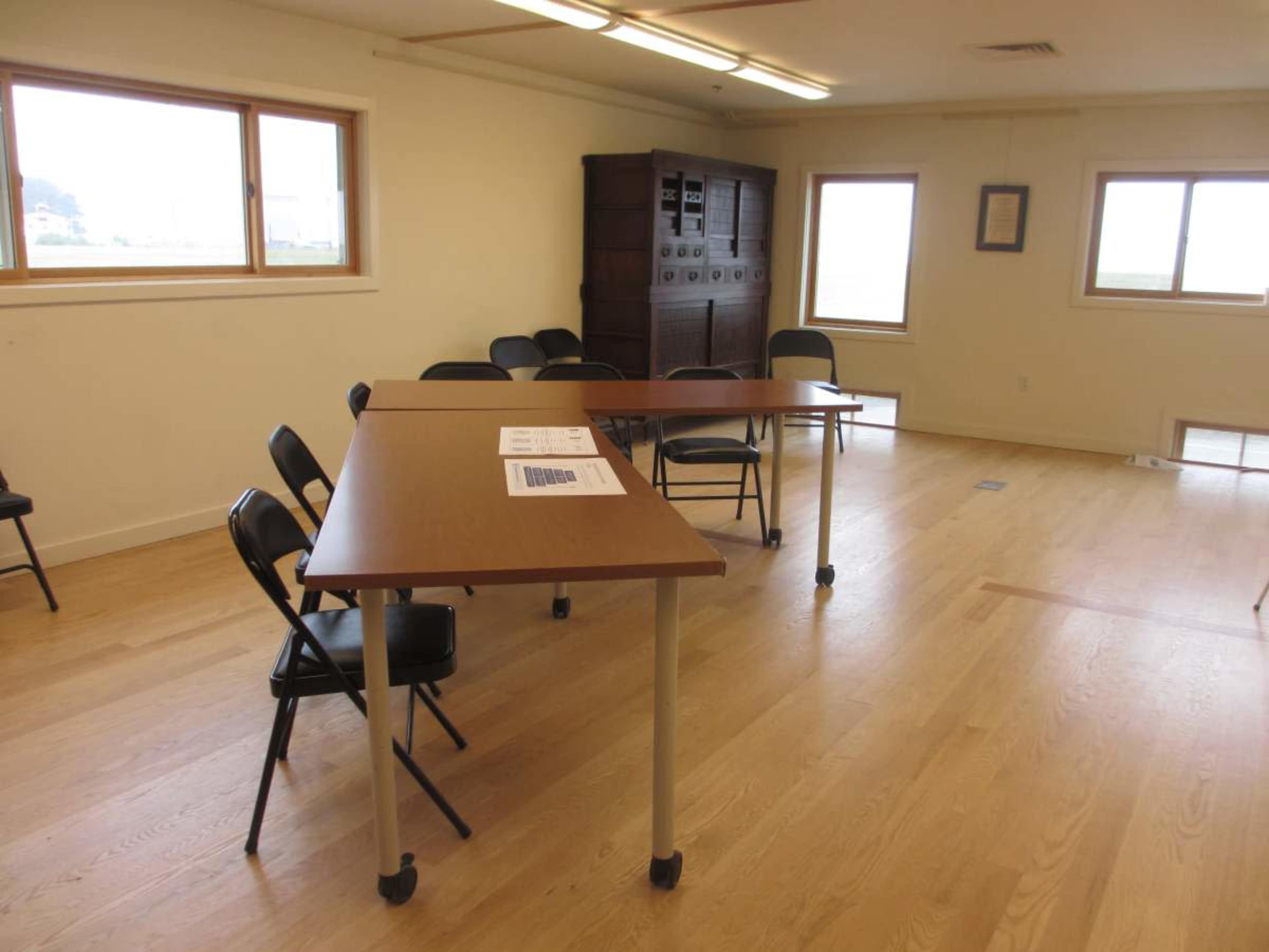 The image shows a simple room with wooden flooring, two tables arranged in an L-shape, and several black chairs placed around them, illuminated by natural light from the windows.
