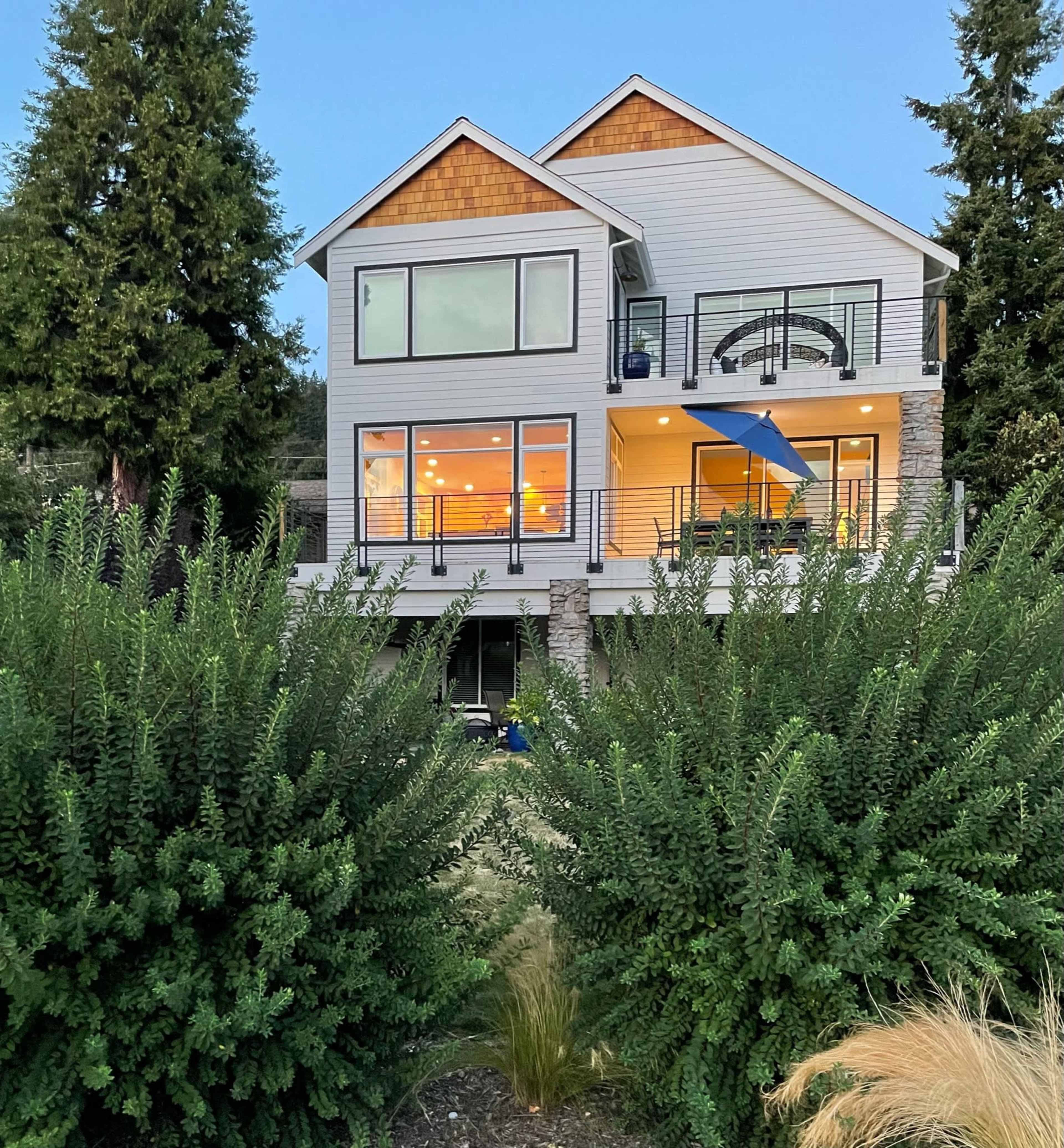 The image shows a modern two-story house with large windows, bright interior lighting, and a balcony, surrounded by lush green shrubs.