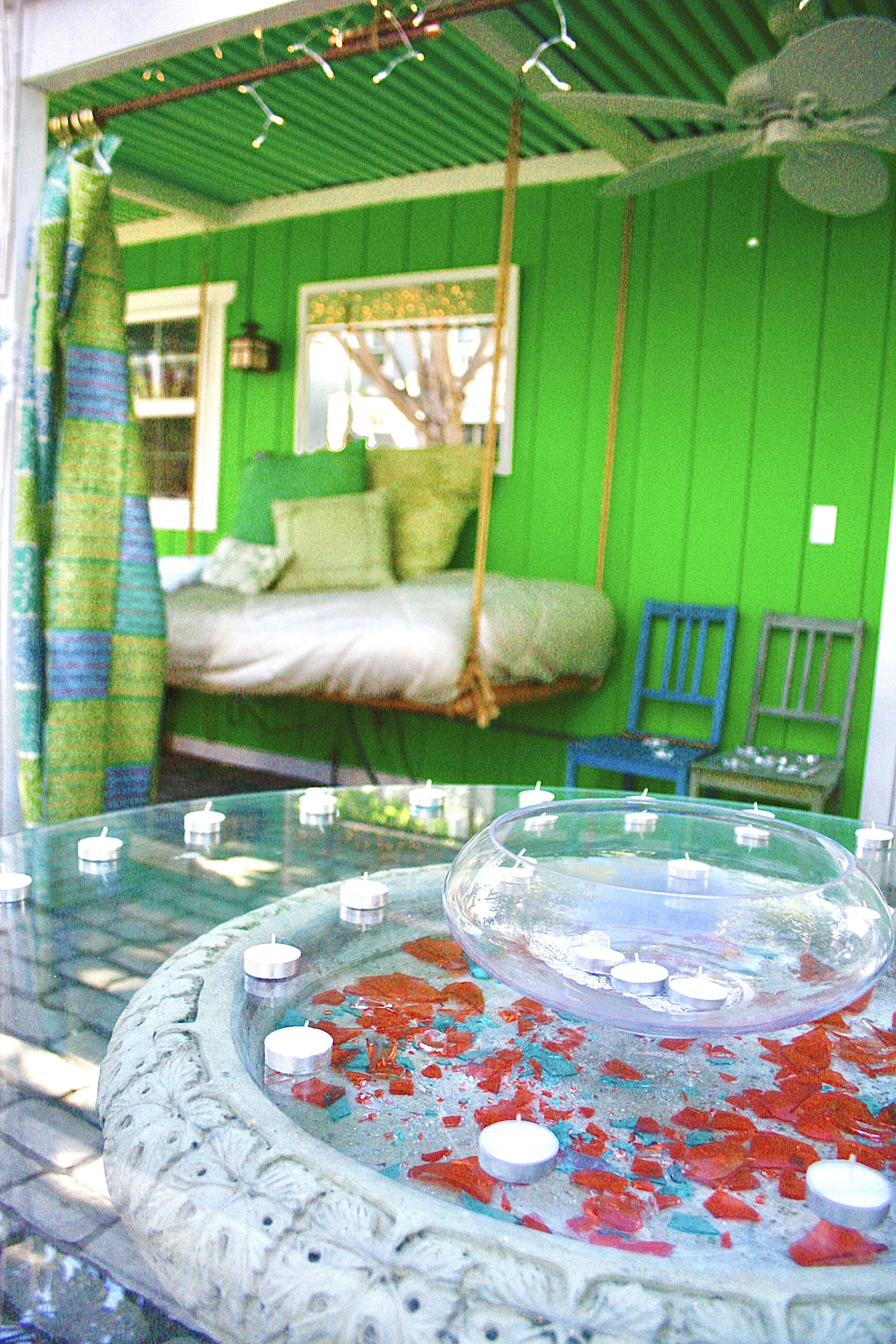 A glass table with scattered red and blue pieces sits in the foreground of a brightly colored green room featuring a hanging bed and blue chairs.