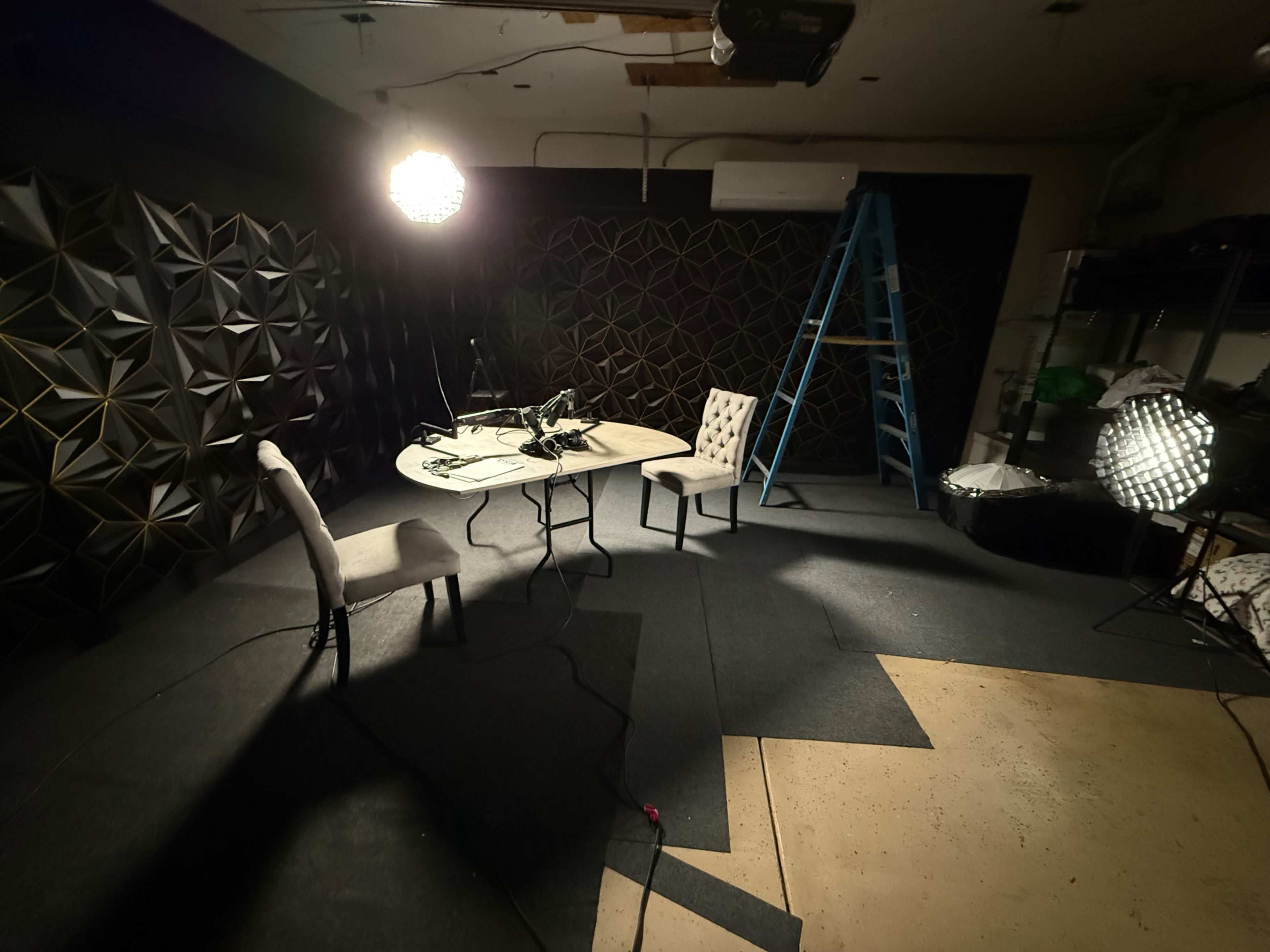 The image shows a dimly lit room with a table and two chairs, a ladder in the corner, and several lighting fixtures set up against a textured black wall.