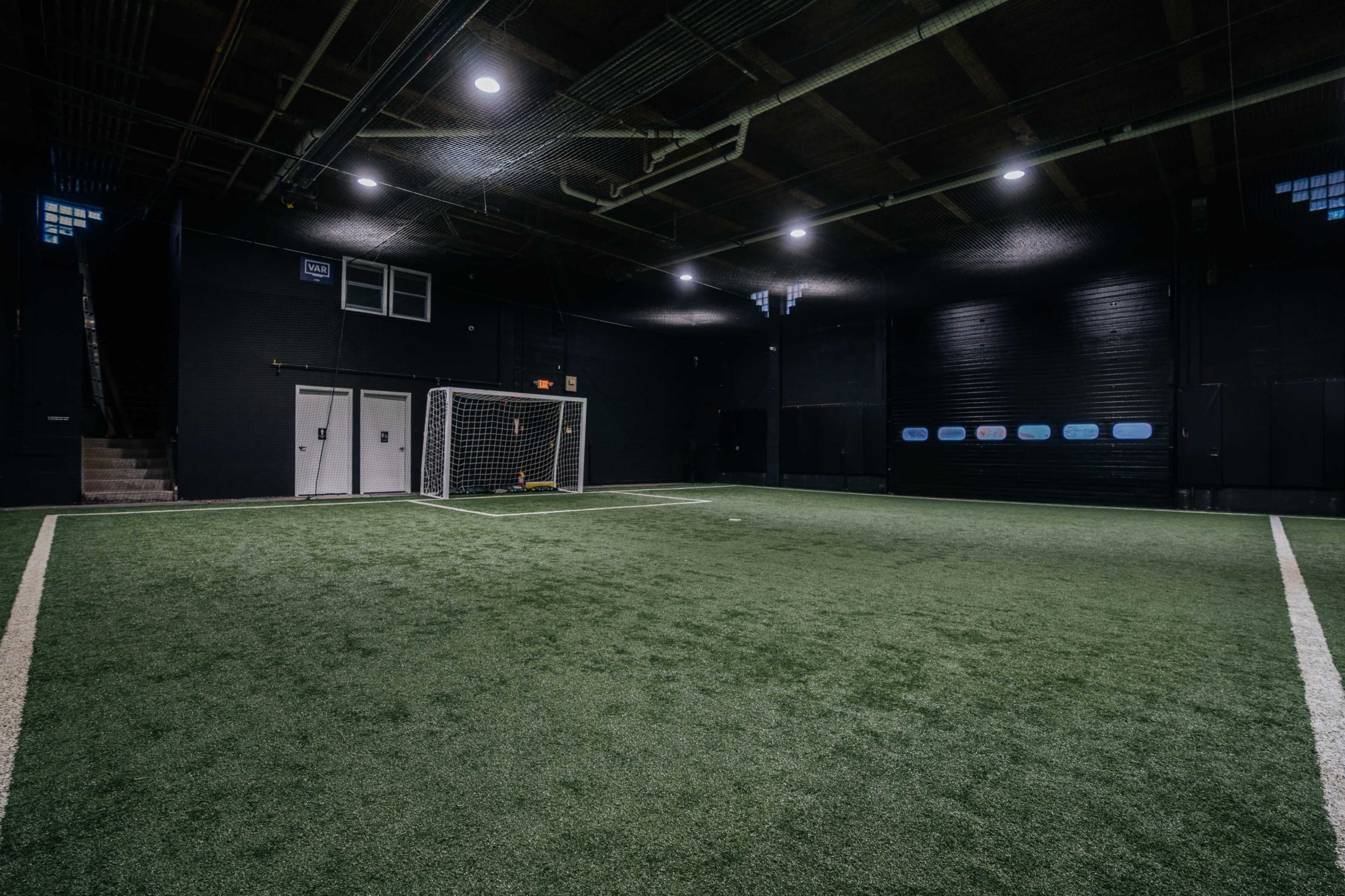 The image shows an indoor soccer field with artificial turf, a goal at one end, and black walls surrounding the space.