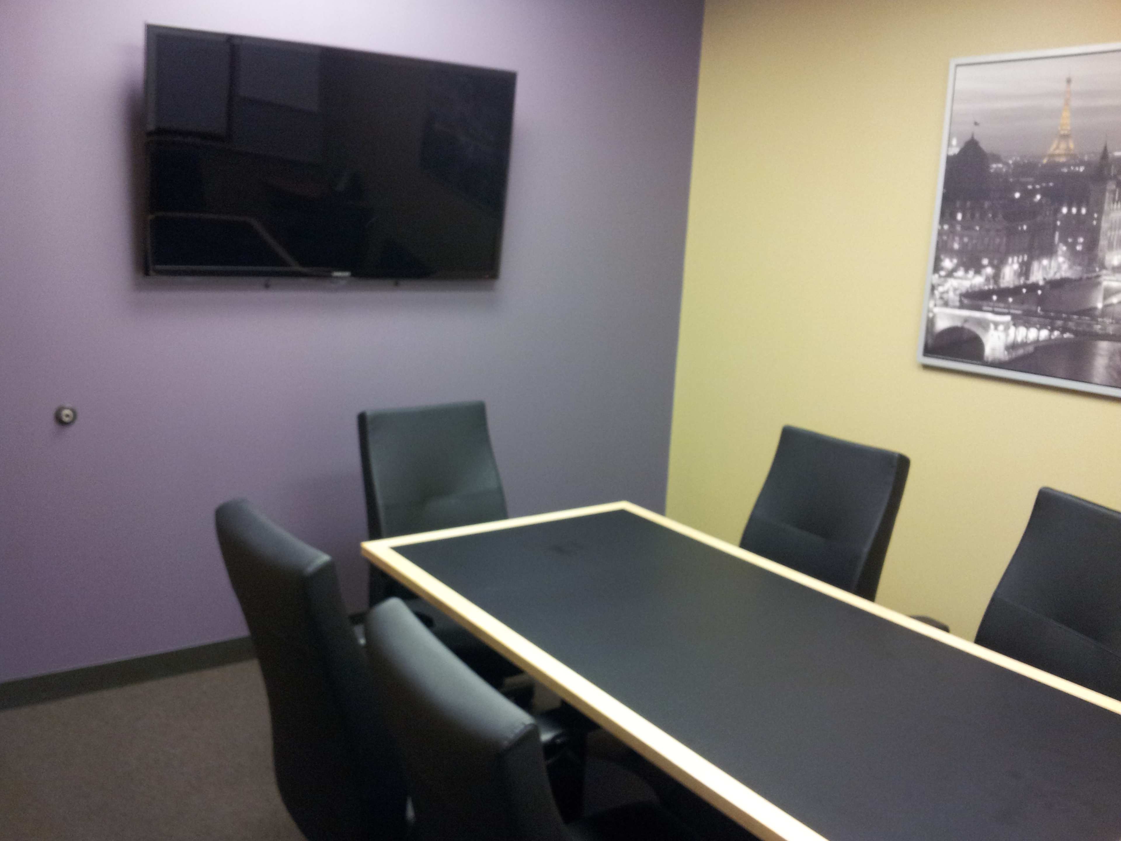 A conference room with a black table, six black chairs, a wall-mounted television, and yellow and purple walls.