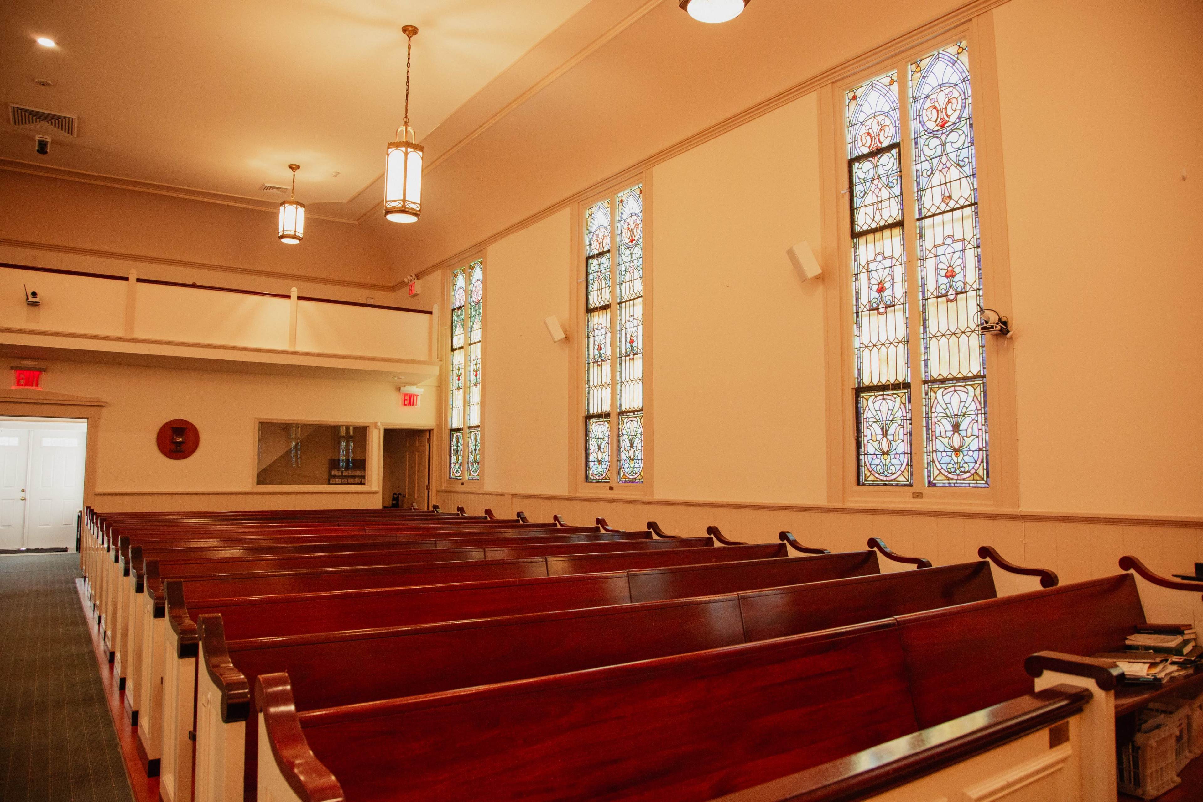 The image shows the interior of a church with wooden pews arranged in rows and stained glass windows along the walls.
