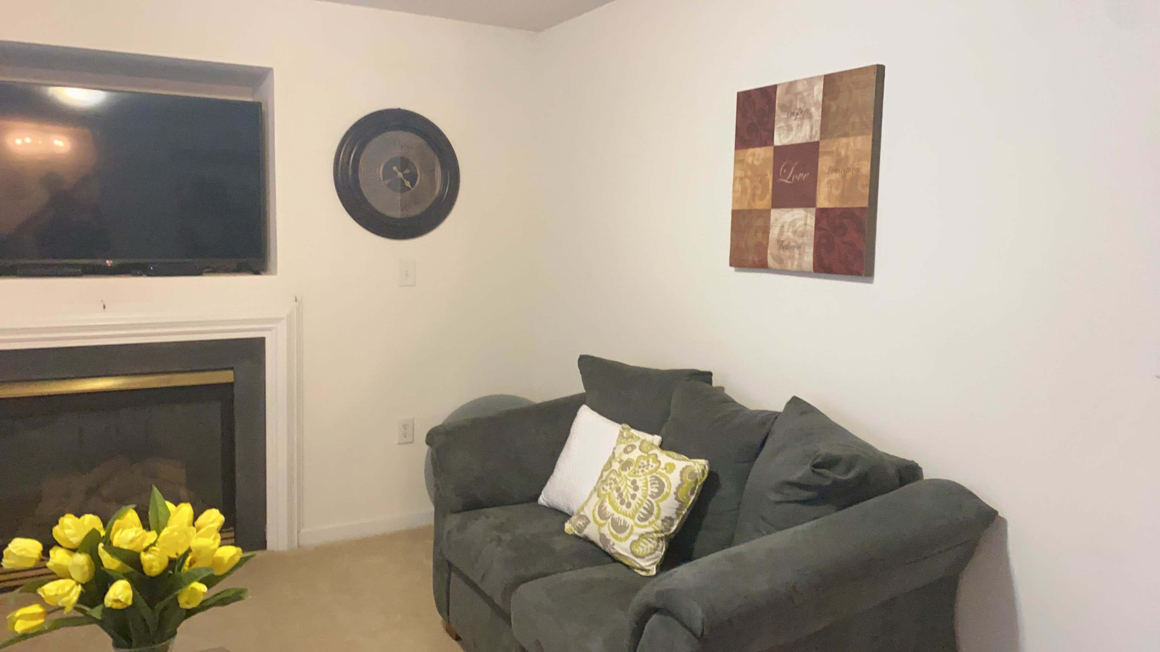 A living room corner with a dark green couch, a decorative wall art piece, a clock, and yellow tulips on a table near a fireplace.
