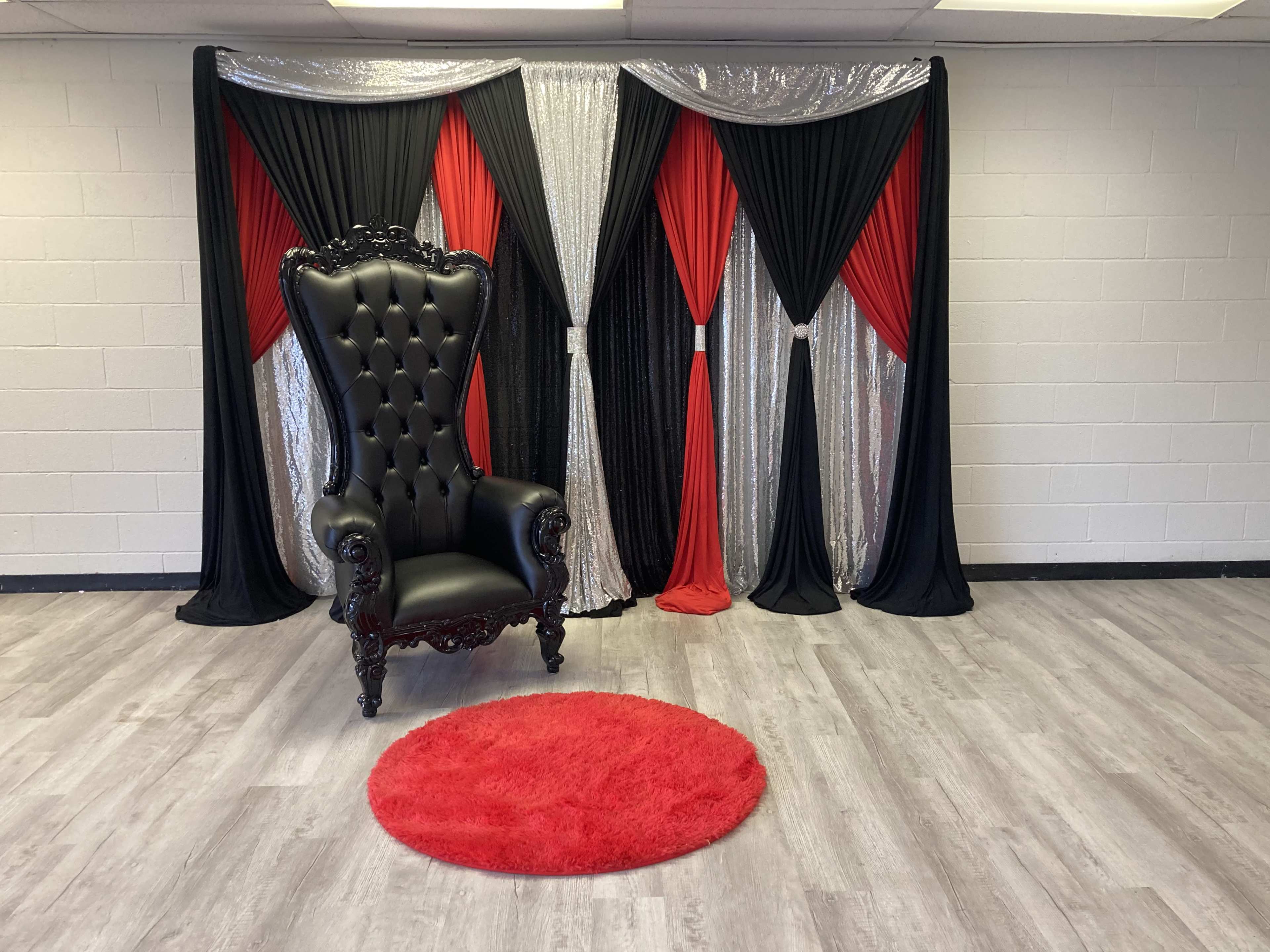 The scene features a black ornate chair in front of a backdrop of red, black, and silver draped fabric, with a red circular rug on the floor.