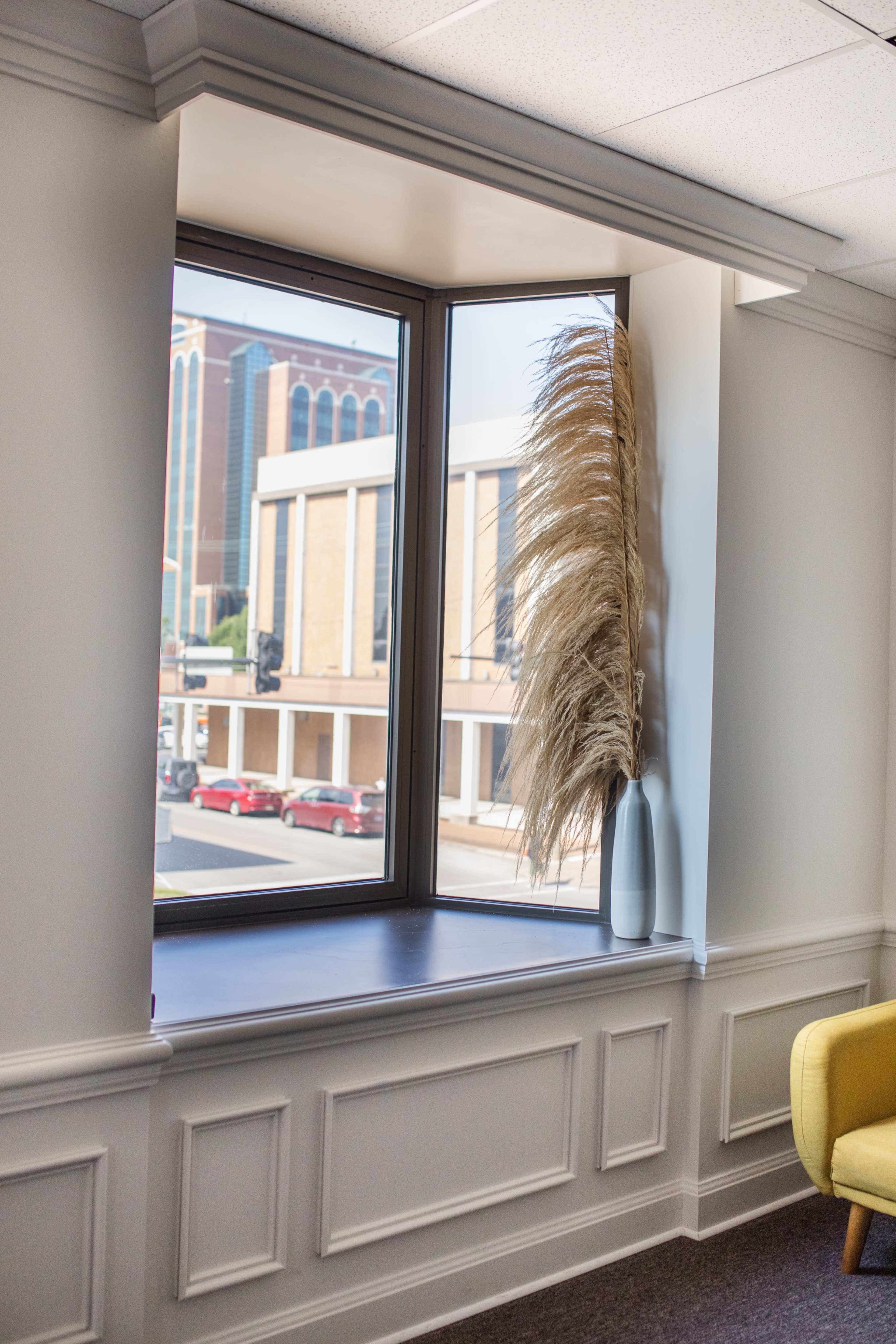 A bright corner window features a tall, dried grass arrangement in a vase, with a view of a city street and buildings outside.