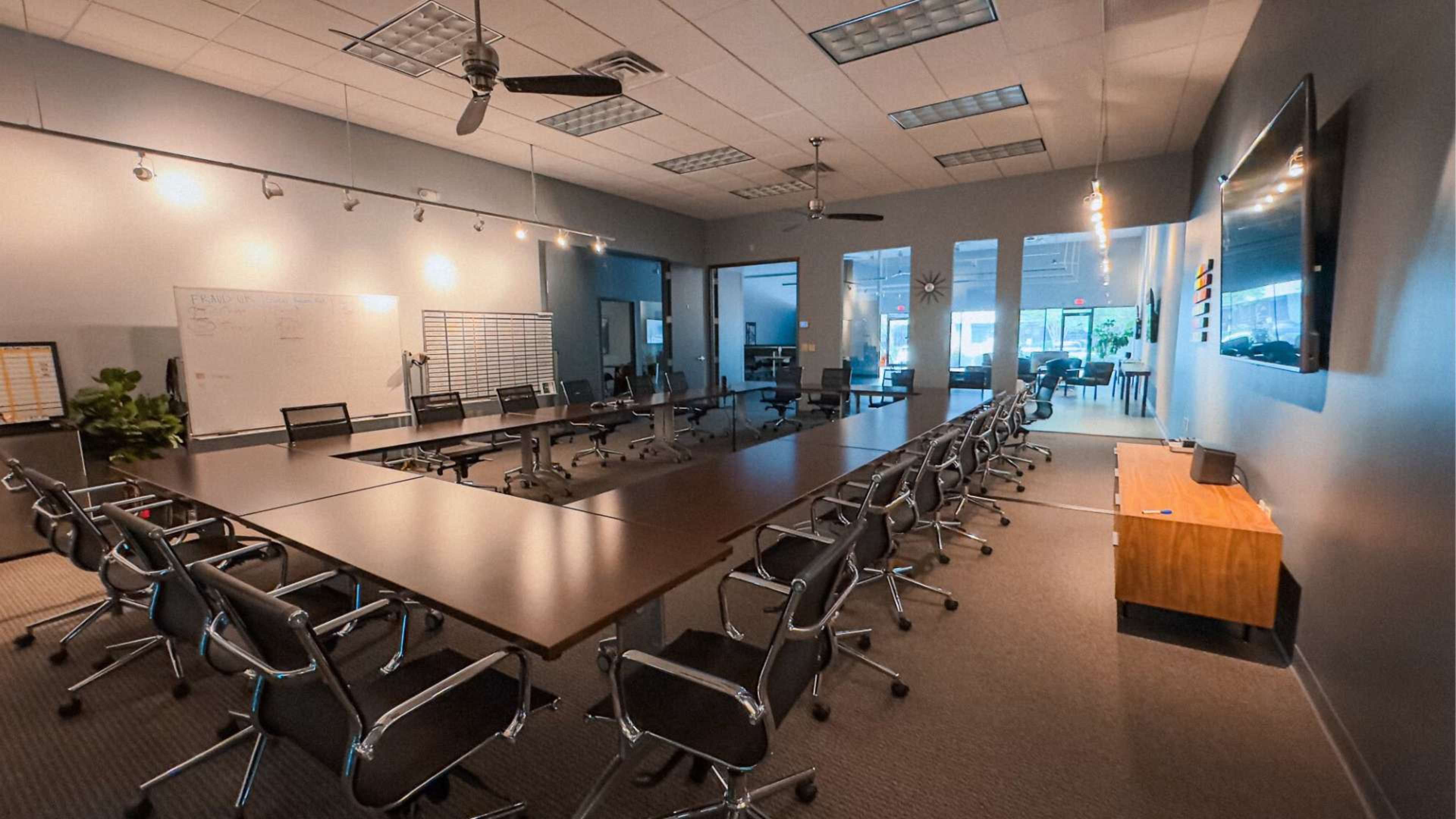A spacious, modern conference room with a long rectangular table surrounded by chairs, along with multiple light sources and a large screen on the wall.