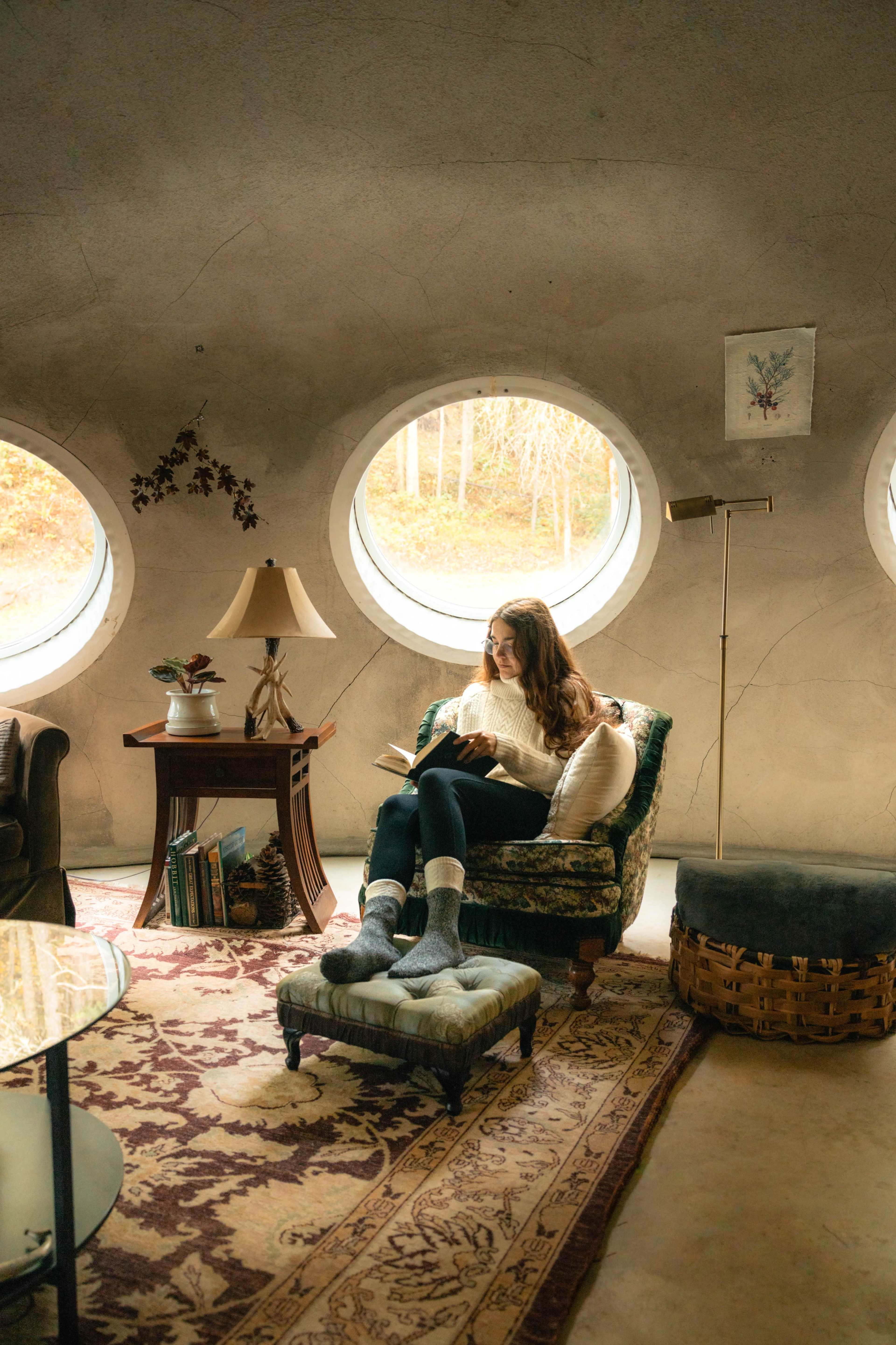 A woman reads a book while seated in an armchair near a circular window in a cozy, modern interior.