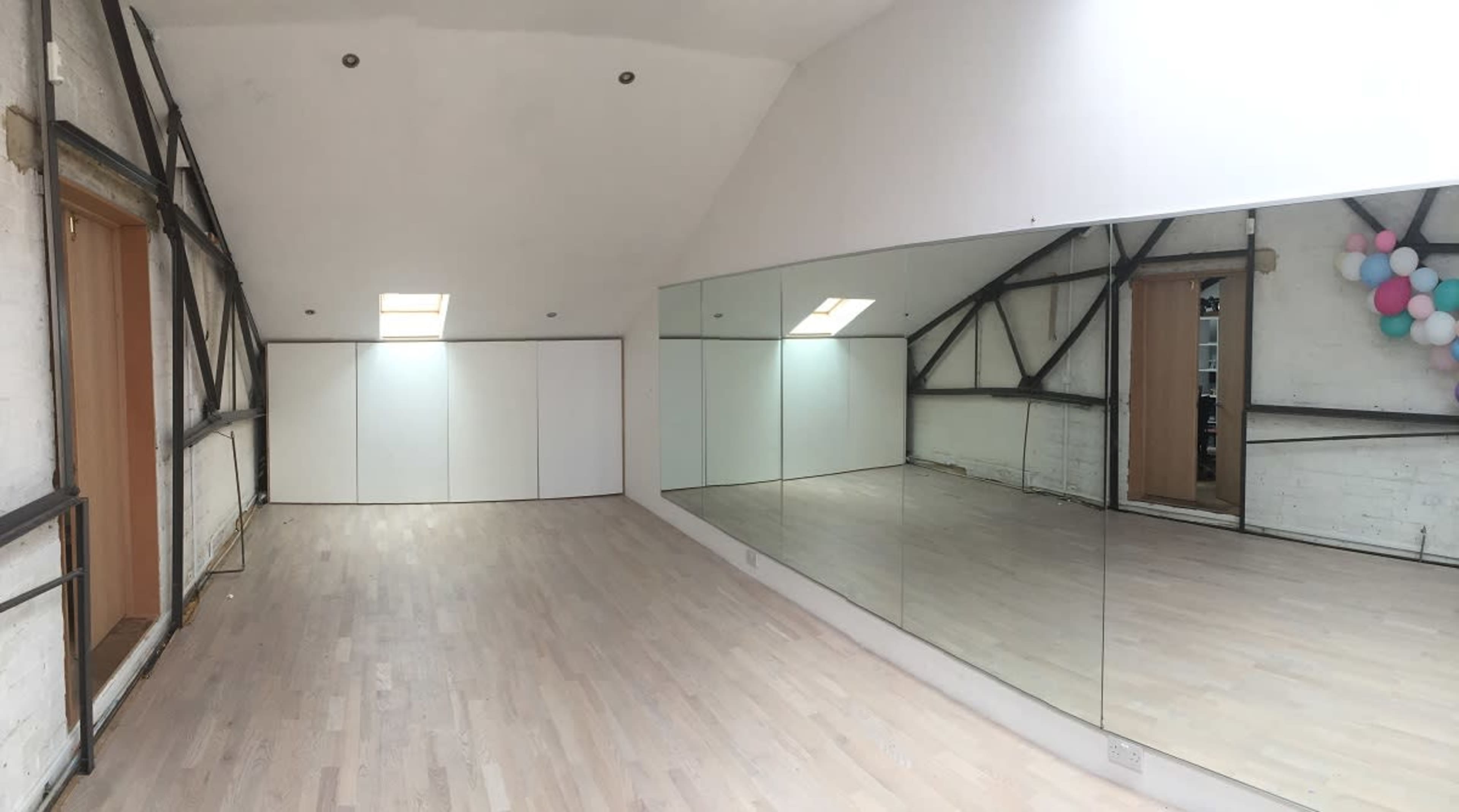 The image shows a spacious room with white walls, wooden flooring, and a large mirror on one side, featuring a door and a small window.