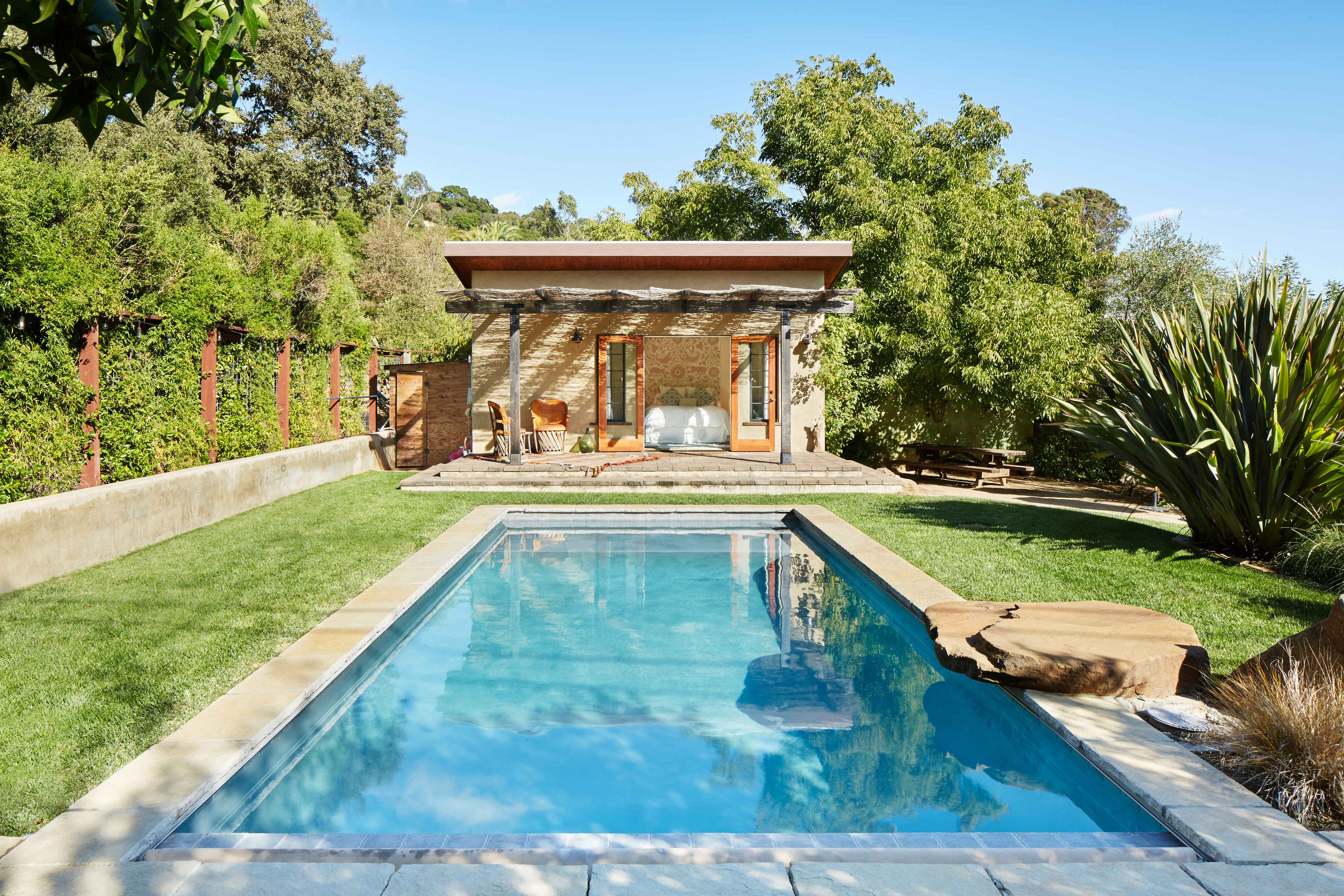 The image shows a modern pool beside a simple, elegant structure surrounded by greenery.
