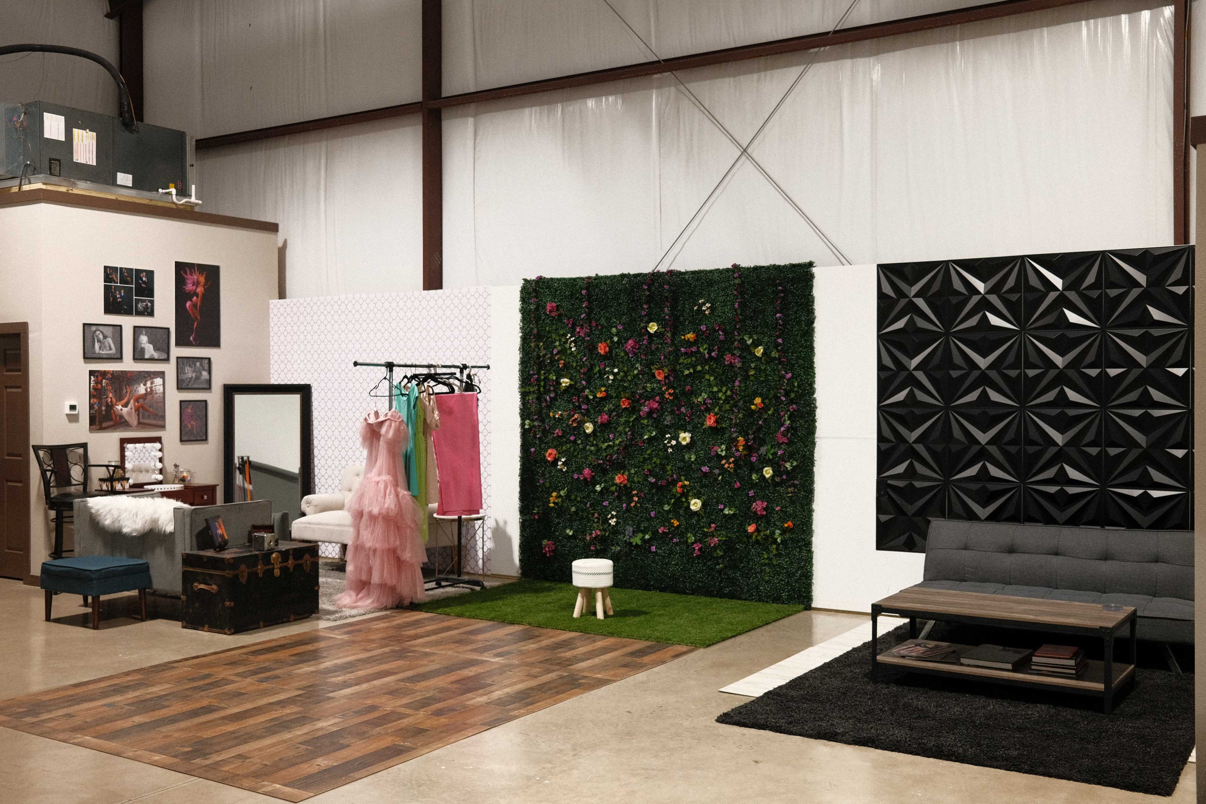 A studio space featuring a mix of furniture including a couch, a dressing area with a mirror, clothing racks, and a decorative wall with greenery and art.