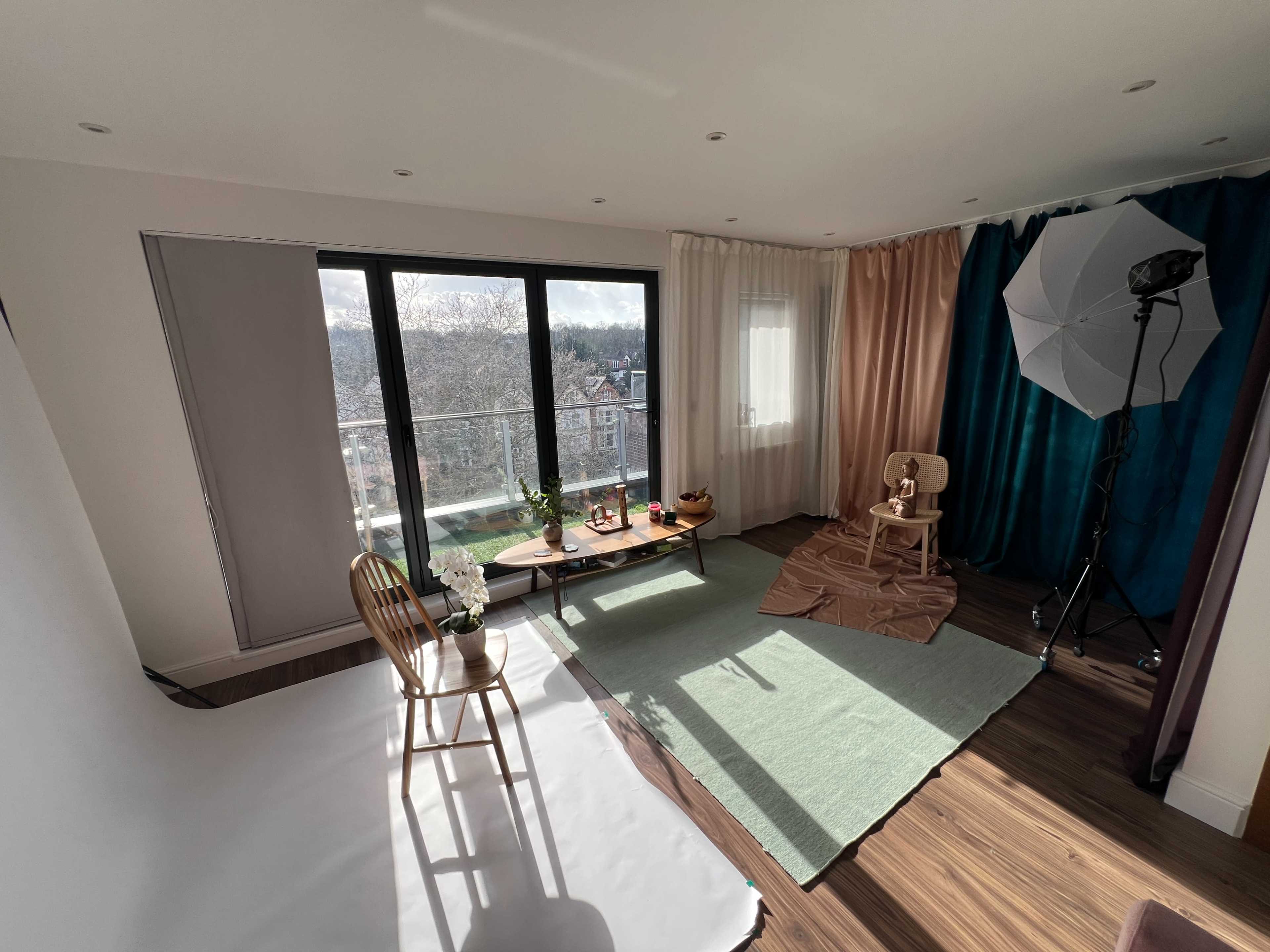 The image shows a bright interior space set up for a photoshoot, featuring a wooden table with several props, a chair, and a backdrop area with different colored curtains.