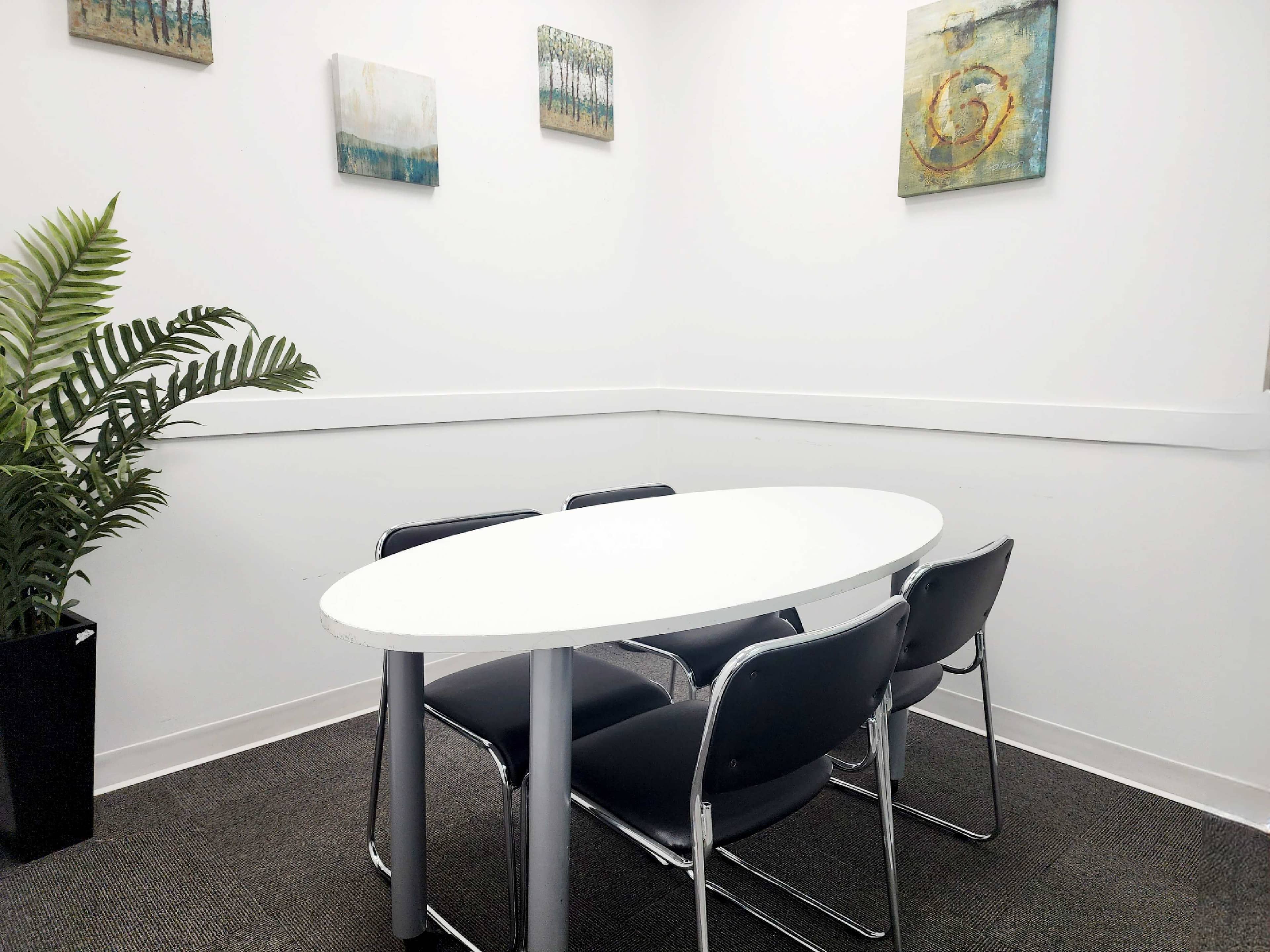 A small meeting room features a white oval table surrounded by four black chairs, with plants and artwork on the walls.