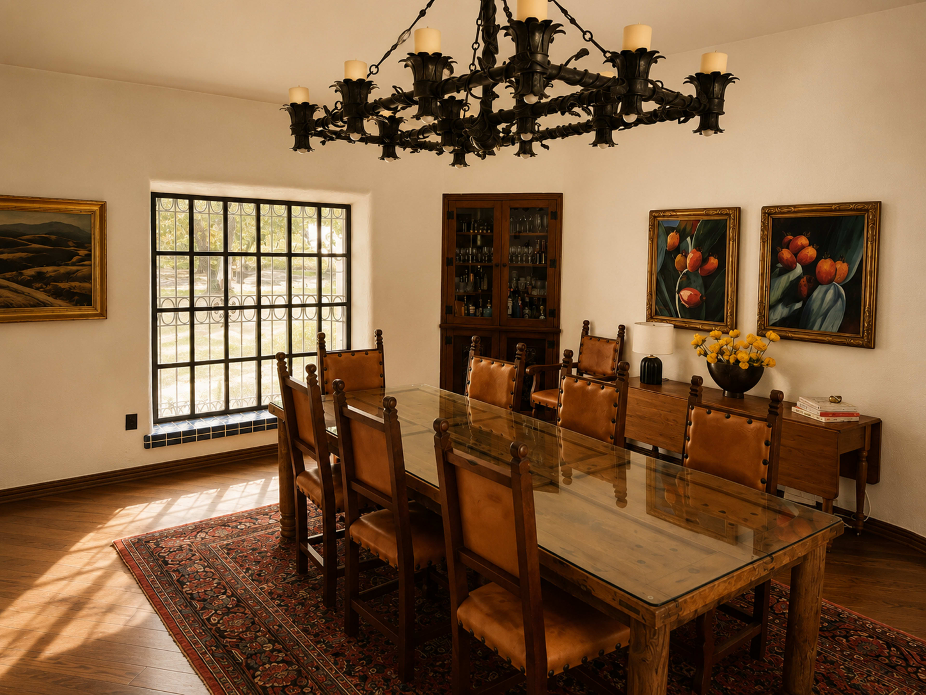 The image shows a dining room featuring a large wooden table with eight chairs, a chandelier overhead, and two paintings on the walls, along with a sideboard and a window providing natural light.