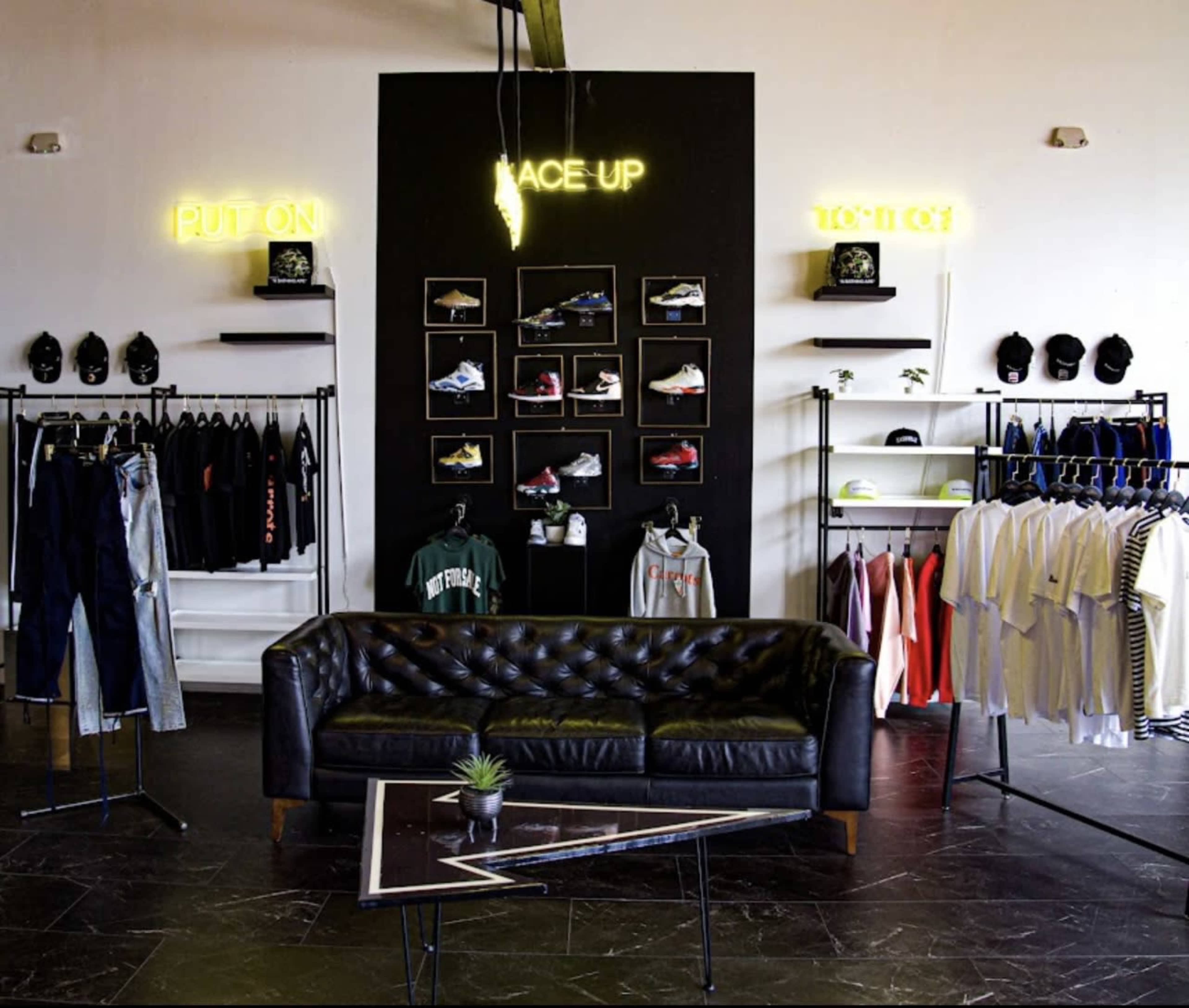 The interior of a retail clothing store features a black wall with neon signs, shelves displaying footwear, and a variety of apparel hanging on racks beside a black leather couch.