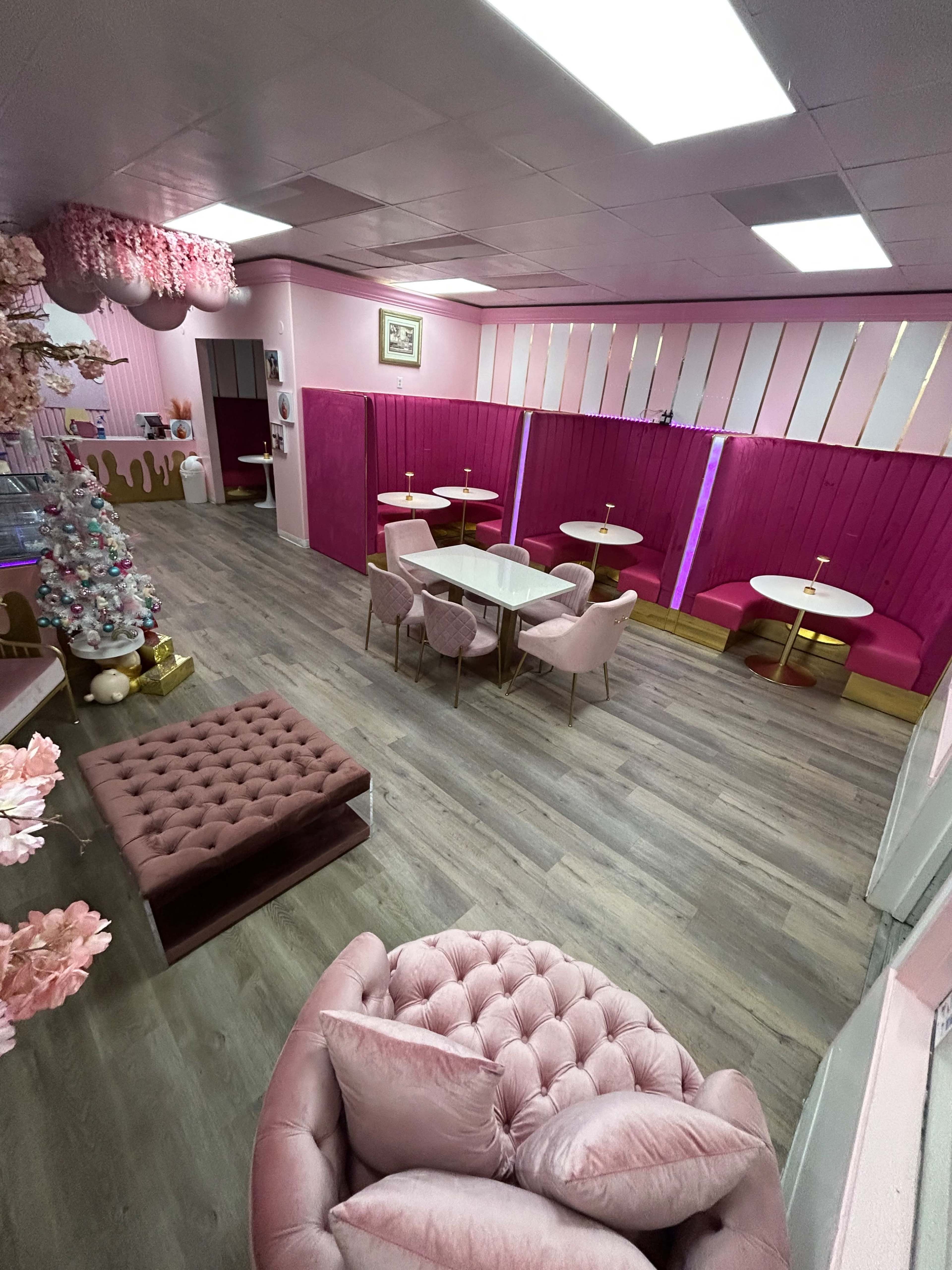 A brightly colored café interior with pink walls, tables surrounded by pink upholstered chairs, and a decorative Christmas tree in the corner.