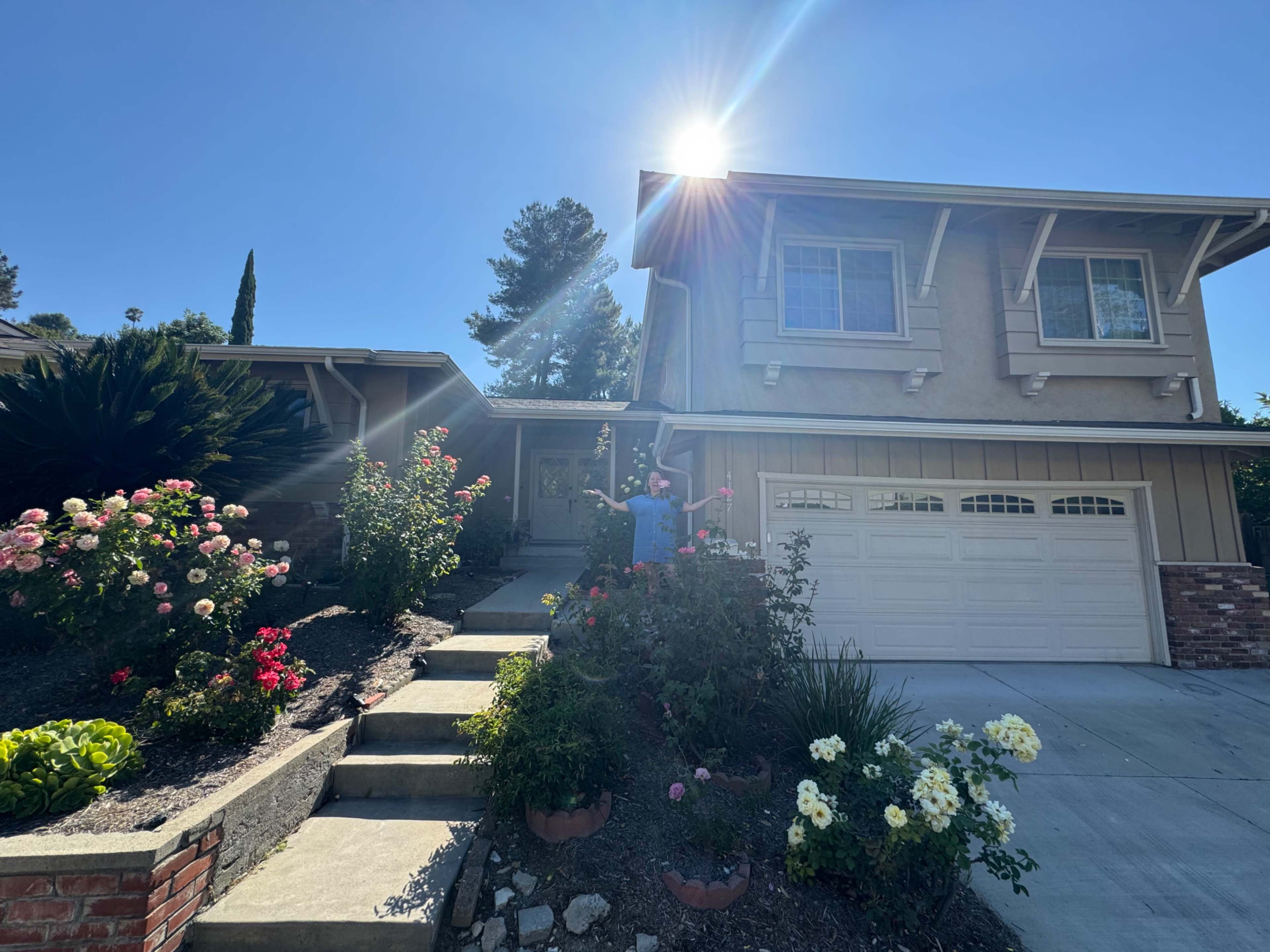 A person stands in front of a two-story house surrounded by flower beds and well-maintained landscaping under a sunny sky.