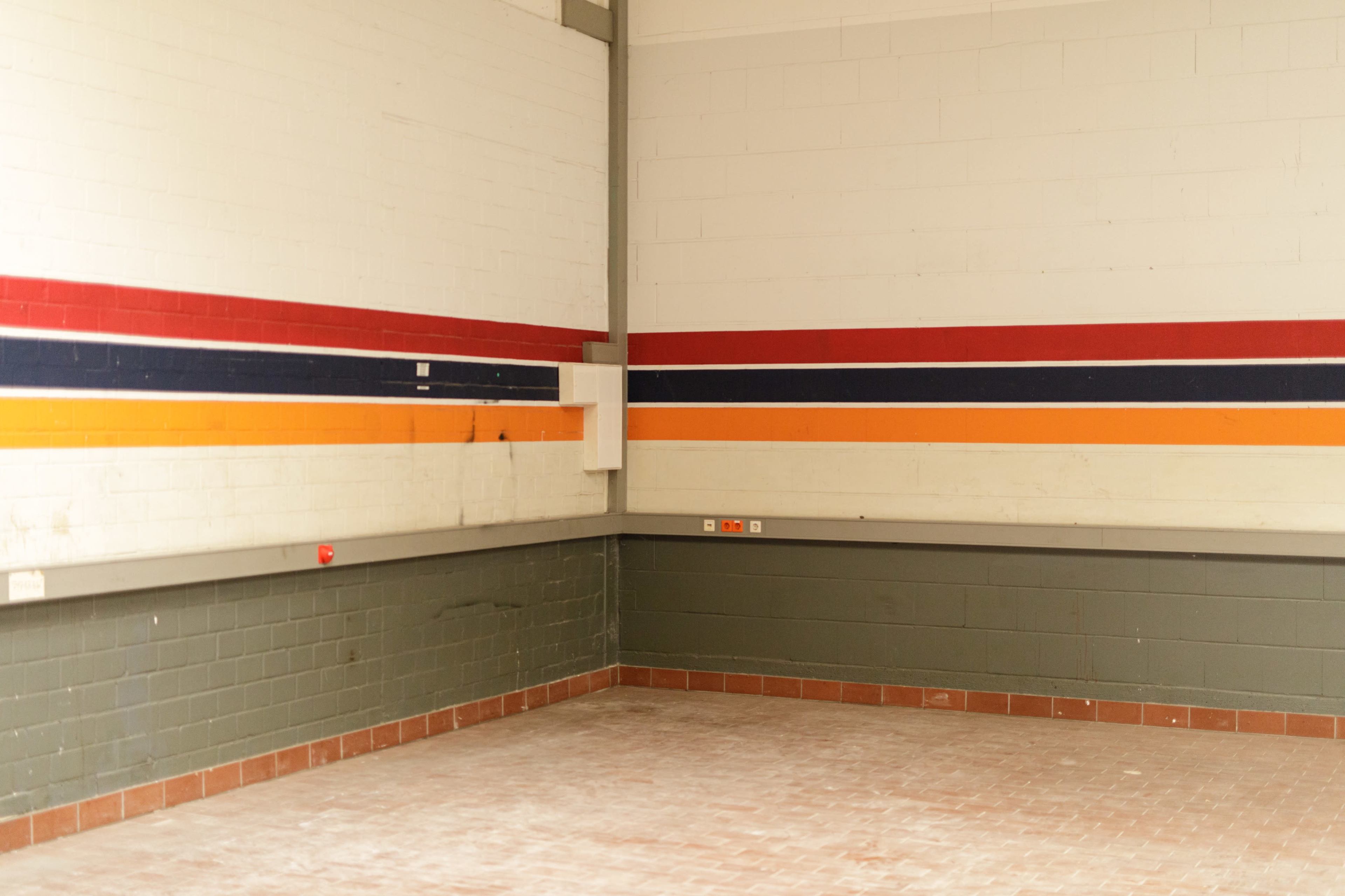 The image shows an empty room with white walls featuring horizontal colored stripes in red, orange, and blue, and a gray tiled floor.