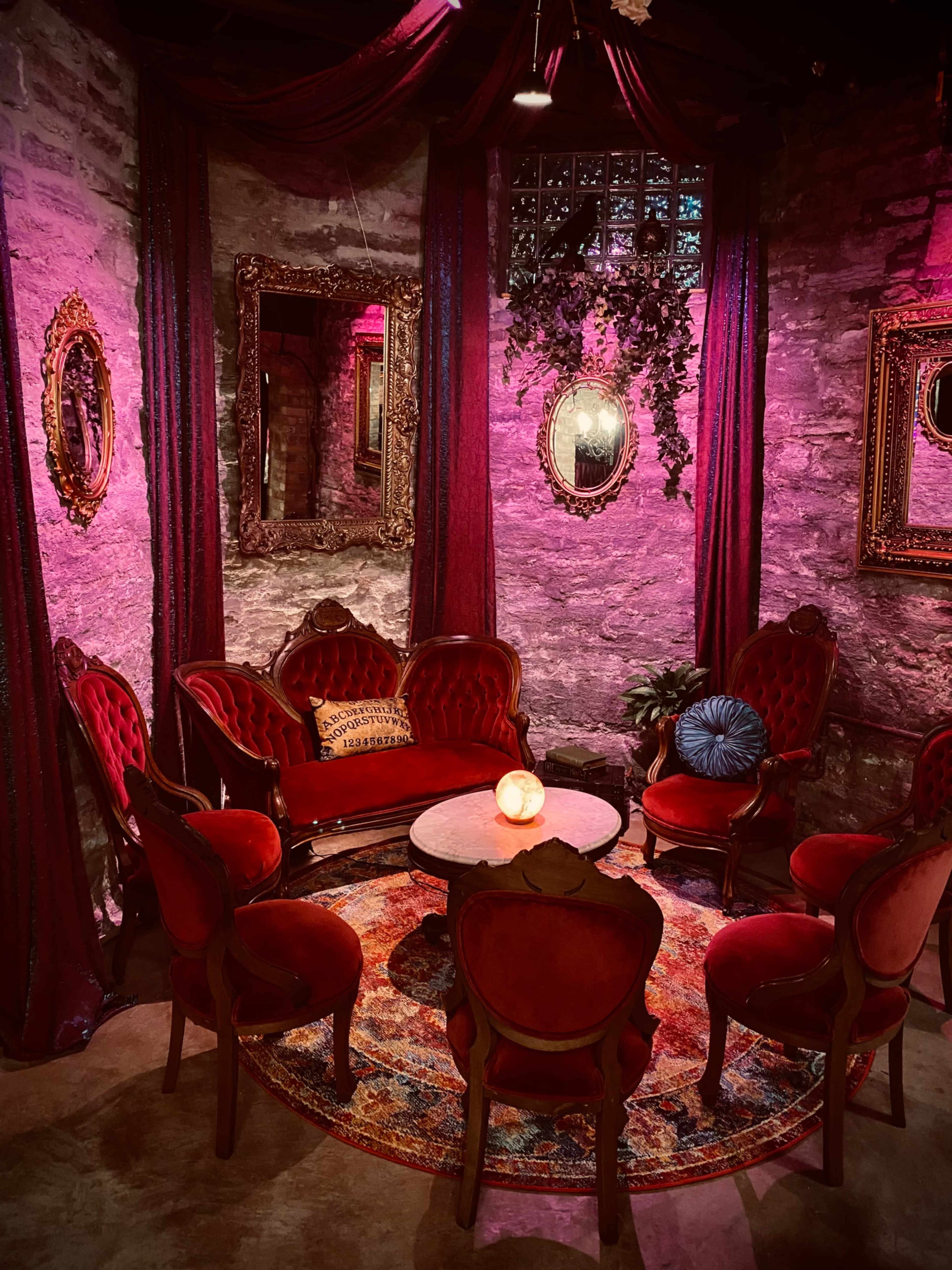 The scene features a cozy seating arrangement with ornate red chairs and a small round table, illuminated by soft lighting in a room with stone walls and decorative mirrors.