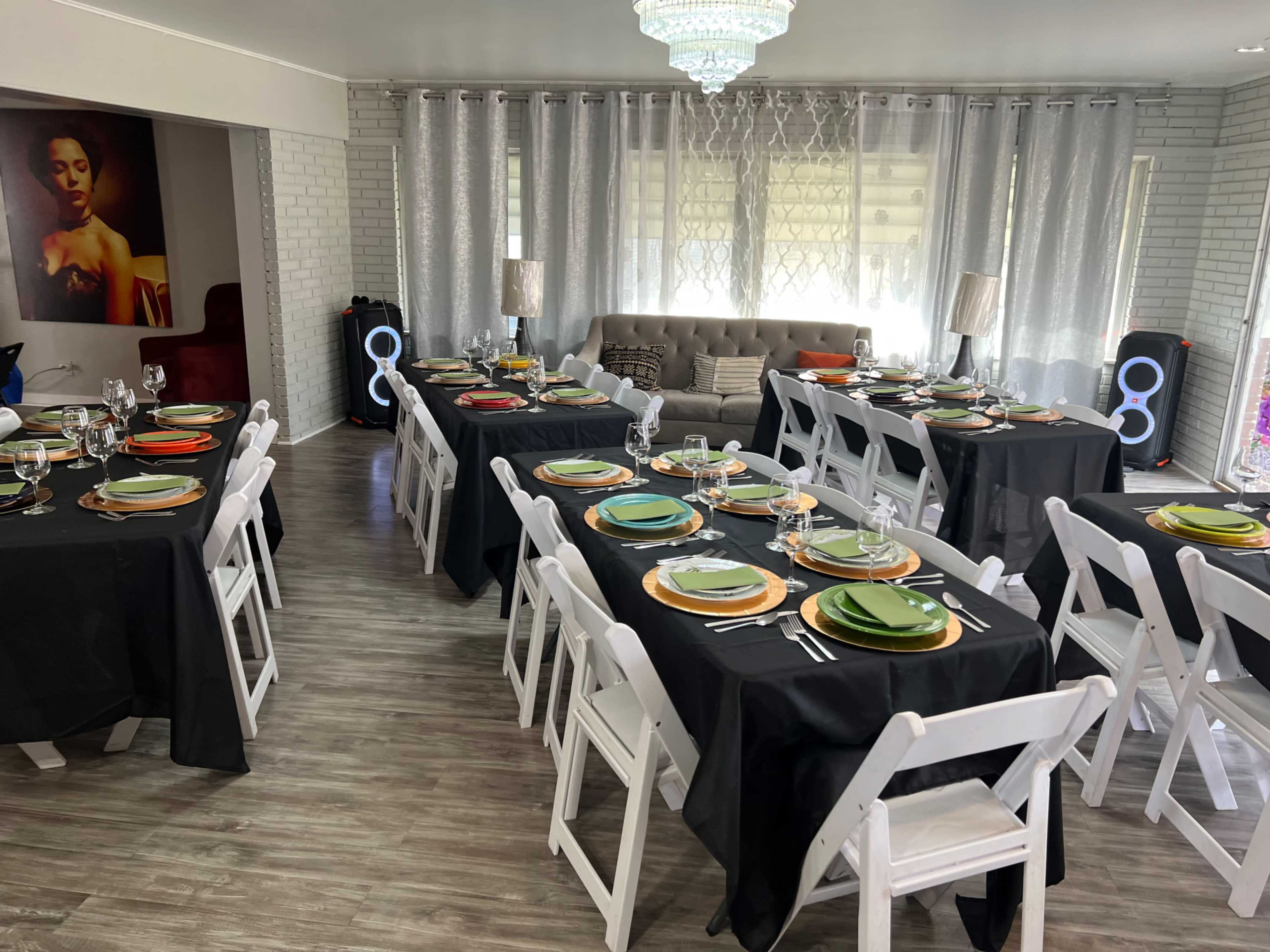 A spacious dining area is set up with multiple tables covered in black cloths, each arranged with colorful plates, utensils, and glasses, alongside a plush sofa and decorative lighting.