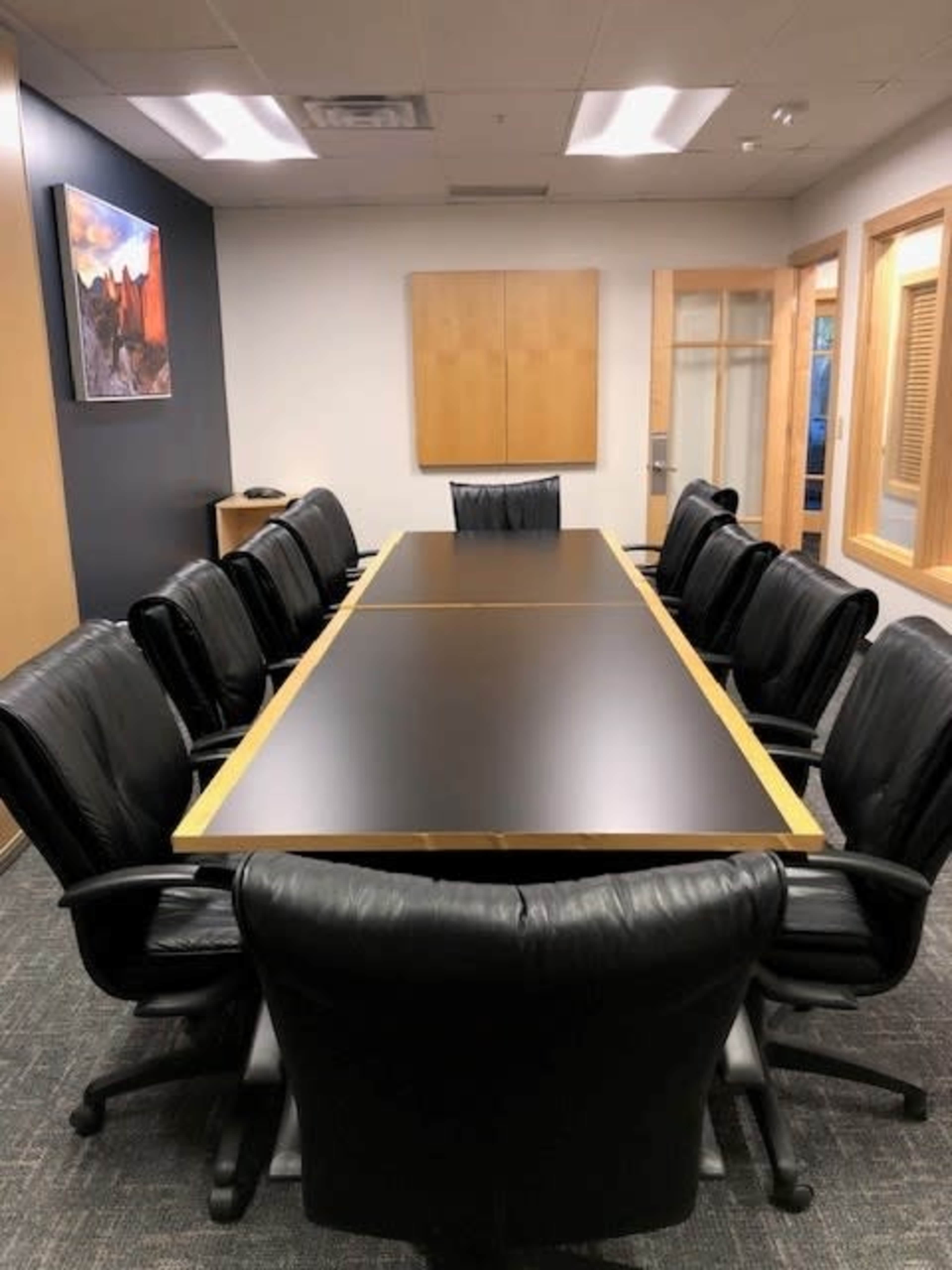 The image depicts a conference room with a long table surrounded by black leather office chairs and a painting on the wall.