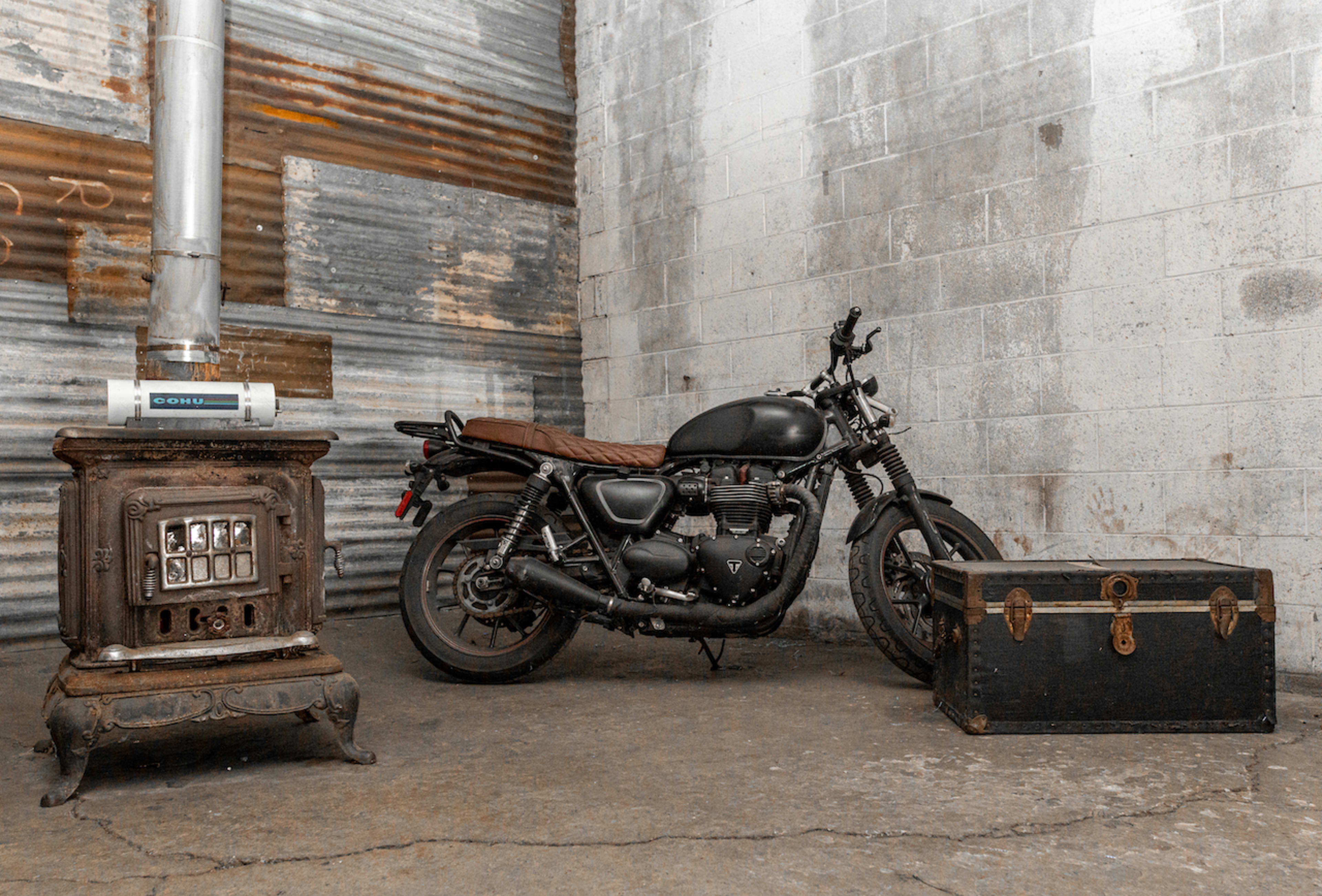 A vintage motorcycle is positioned next to an old trunk and a metal stove in a weathered industrial space.