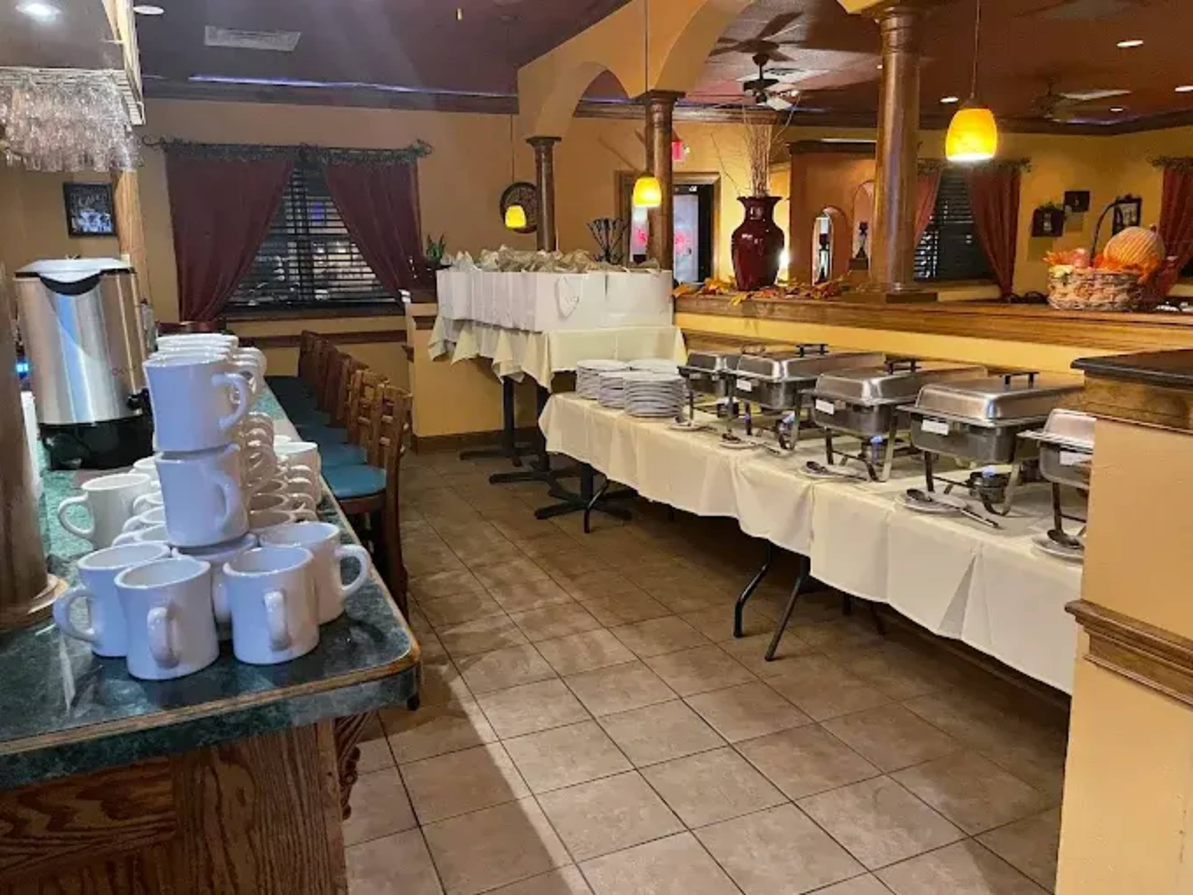 The image shows a restaurant interior with a row of covered food trays on tables, stacked plates, and a coffee station with mugs arranged nearby.