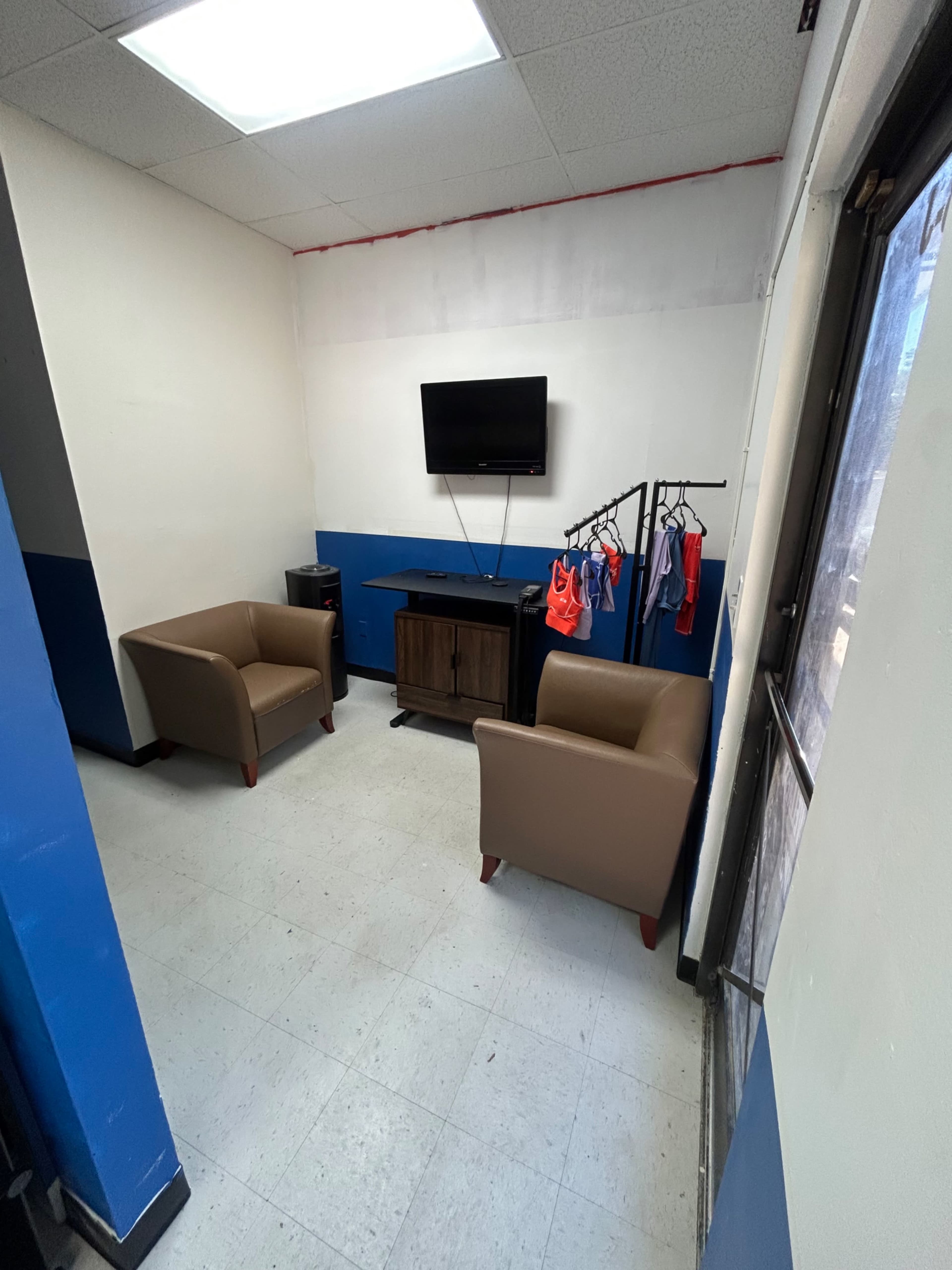 The image shows a small waiting area featuring two brown armchairs, a television mounted on the wall, and a clothing rack with hung apparel.