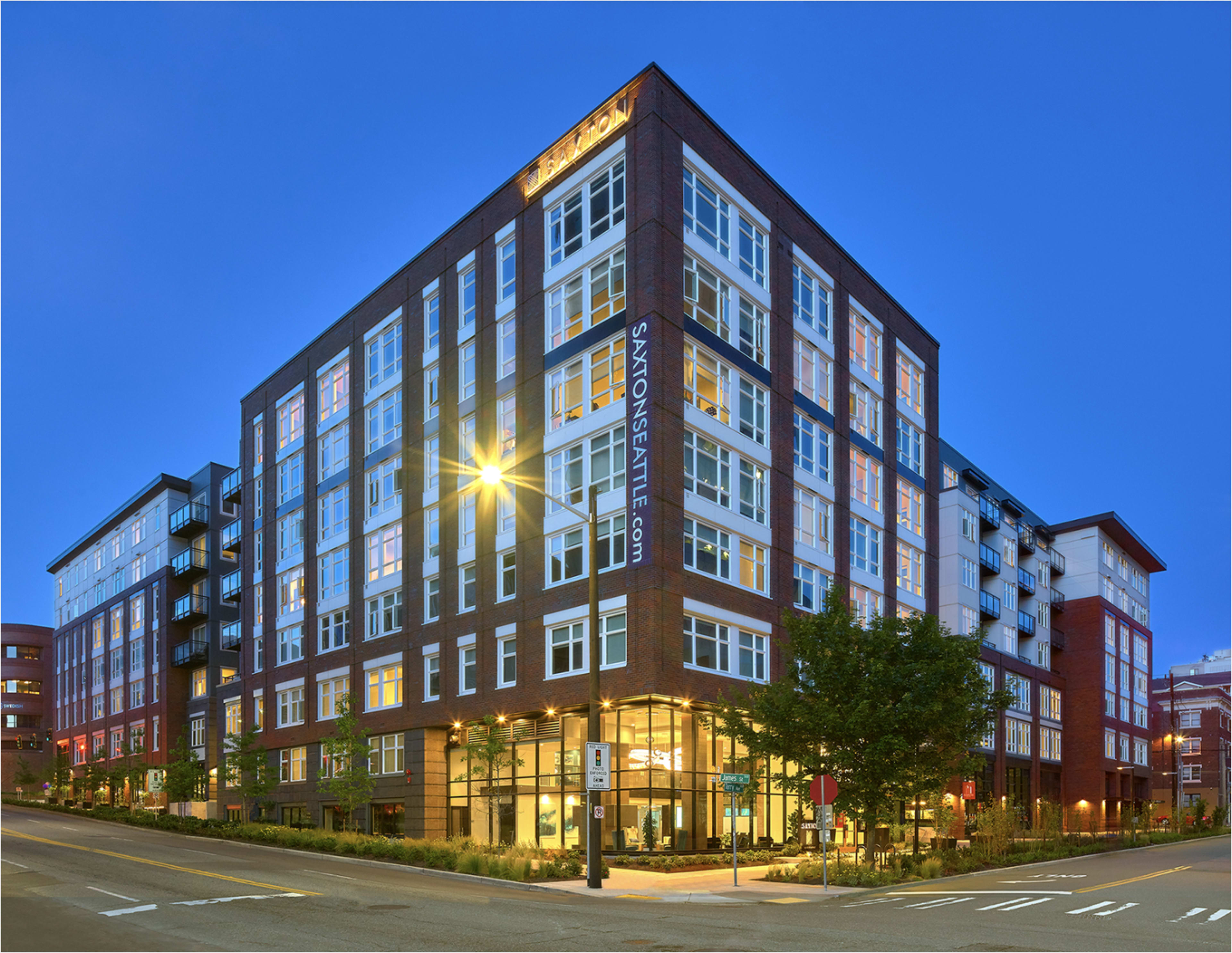 The image shows a modern, multi-story brick apartment building with large windows and street-level signage, illuminated in the evening light.