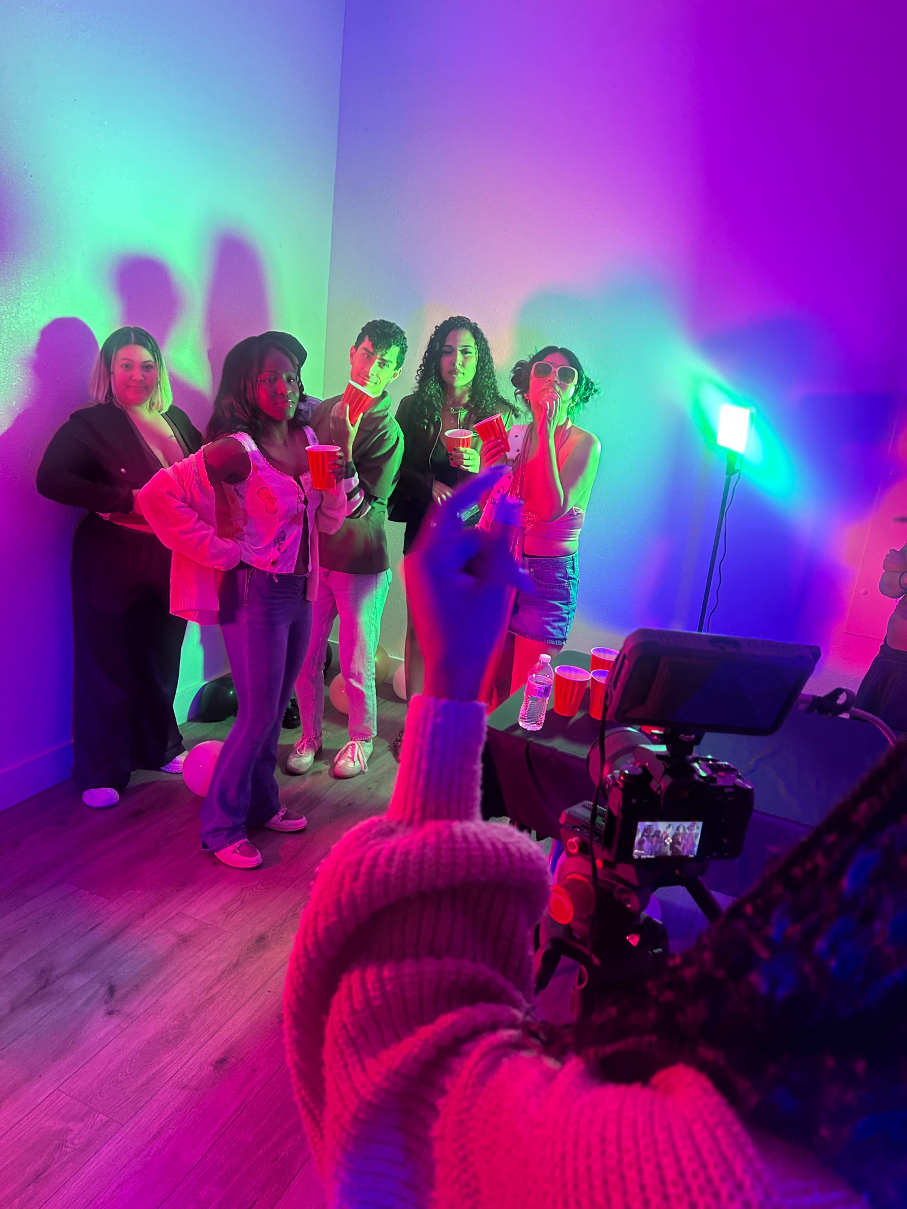 A group of five people stands against a brightly lit wall, holding drinks and posing for a photo while a person in the foreground captures the moment with a camera.