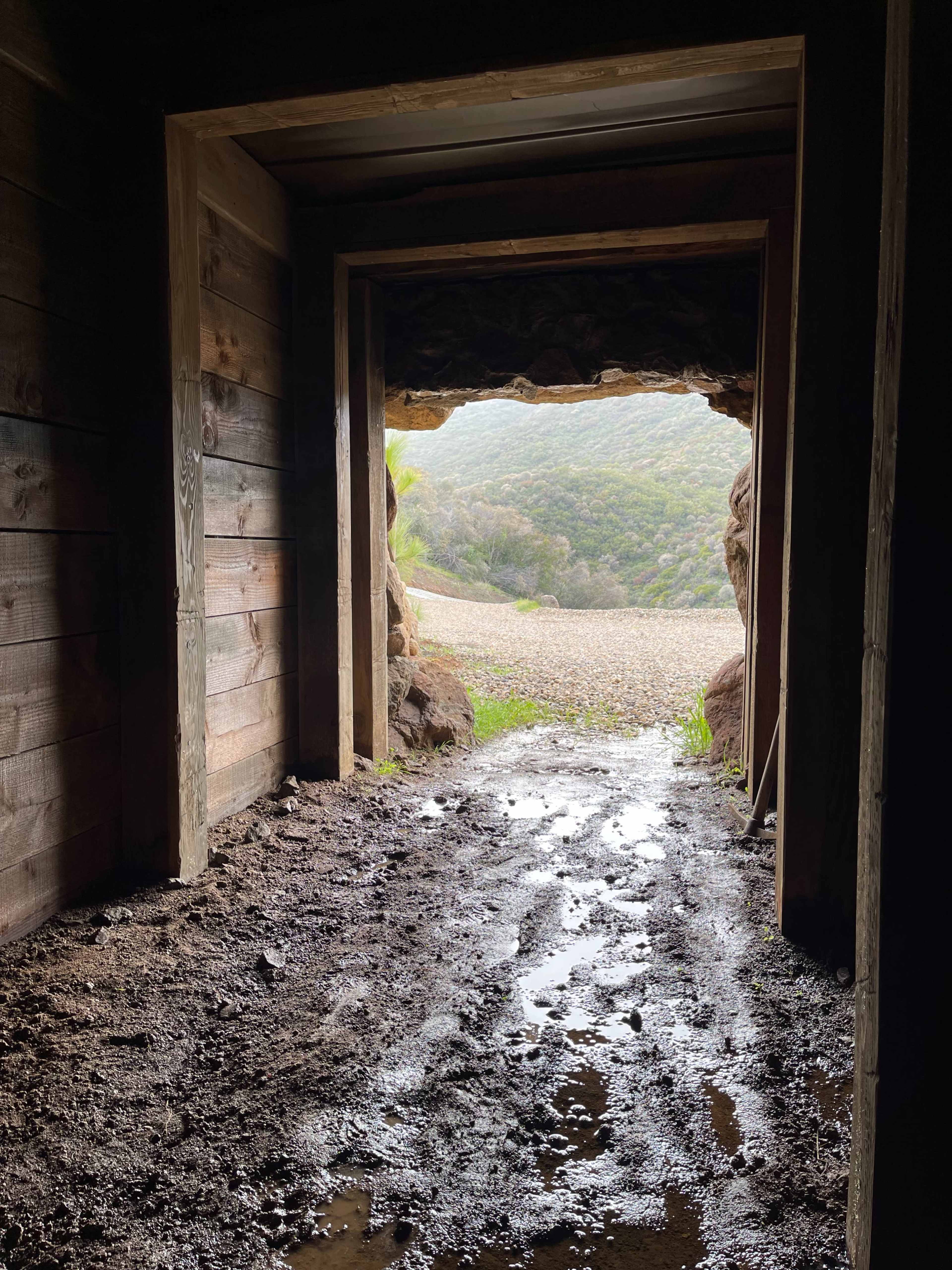 The image shows the view from inside a wooden structure looking out through a rocky opening onto a landscape with green hills and a gravel path.