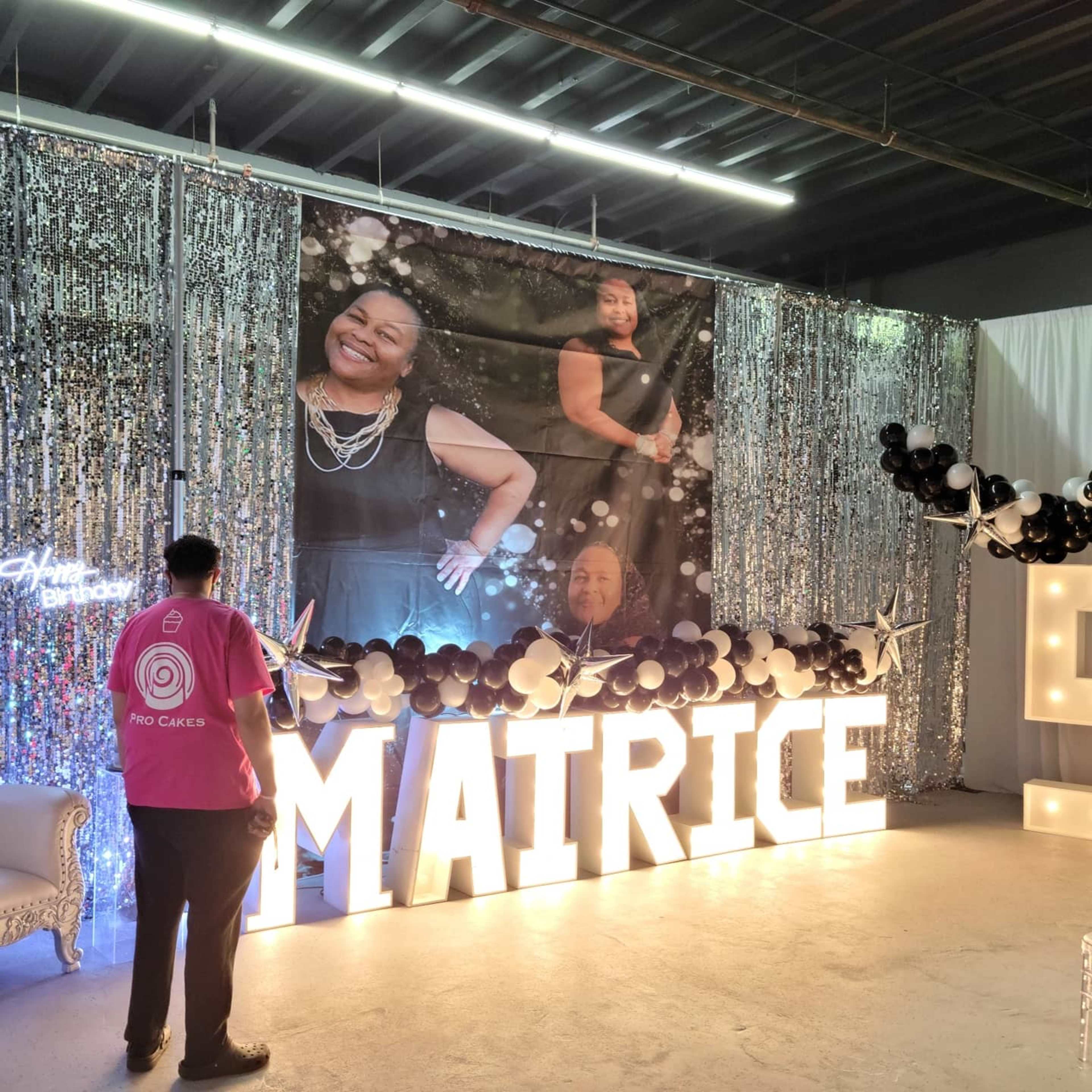 A person stands in front of a decorative backdrop featuring a large photo of an individual, with illuminated letters spelling out "MAITRICE" and sparkling curtains.