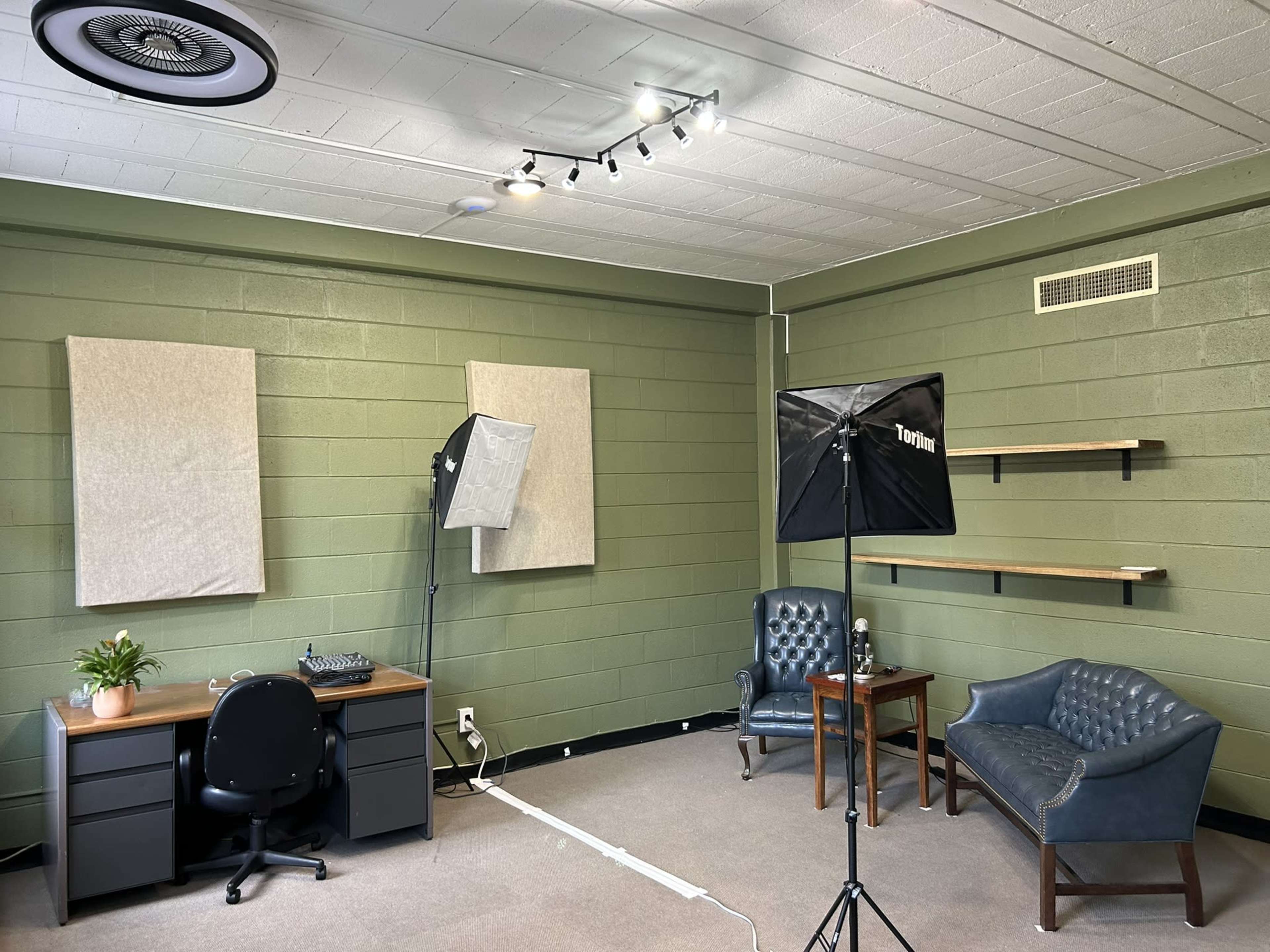 The image shows a sparsely furnished room with green walls, a desk with audio equipment, two armchairs, and a small wooden table.