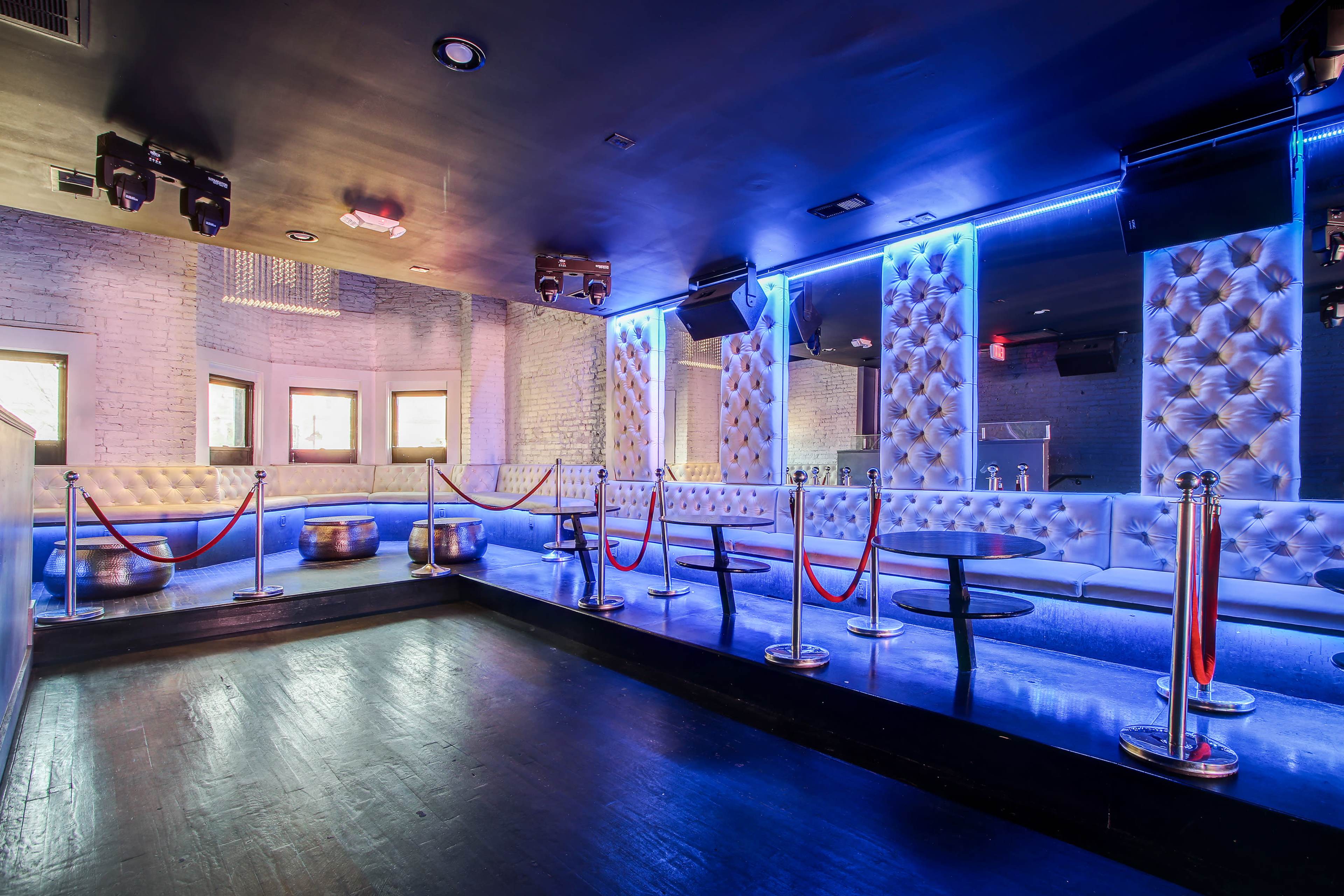 A modern lounge area with patterned seating, illuminated by blue neon lights and featuring a dance floor separated by red velvet ropes.