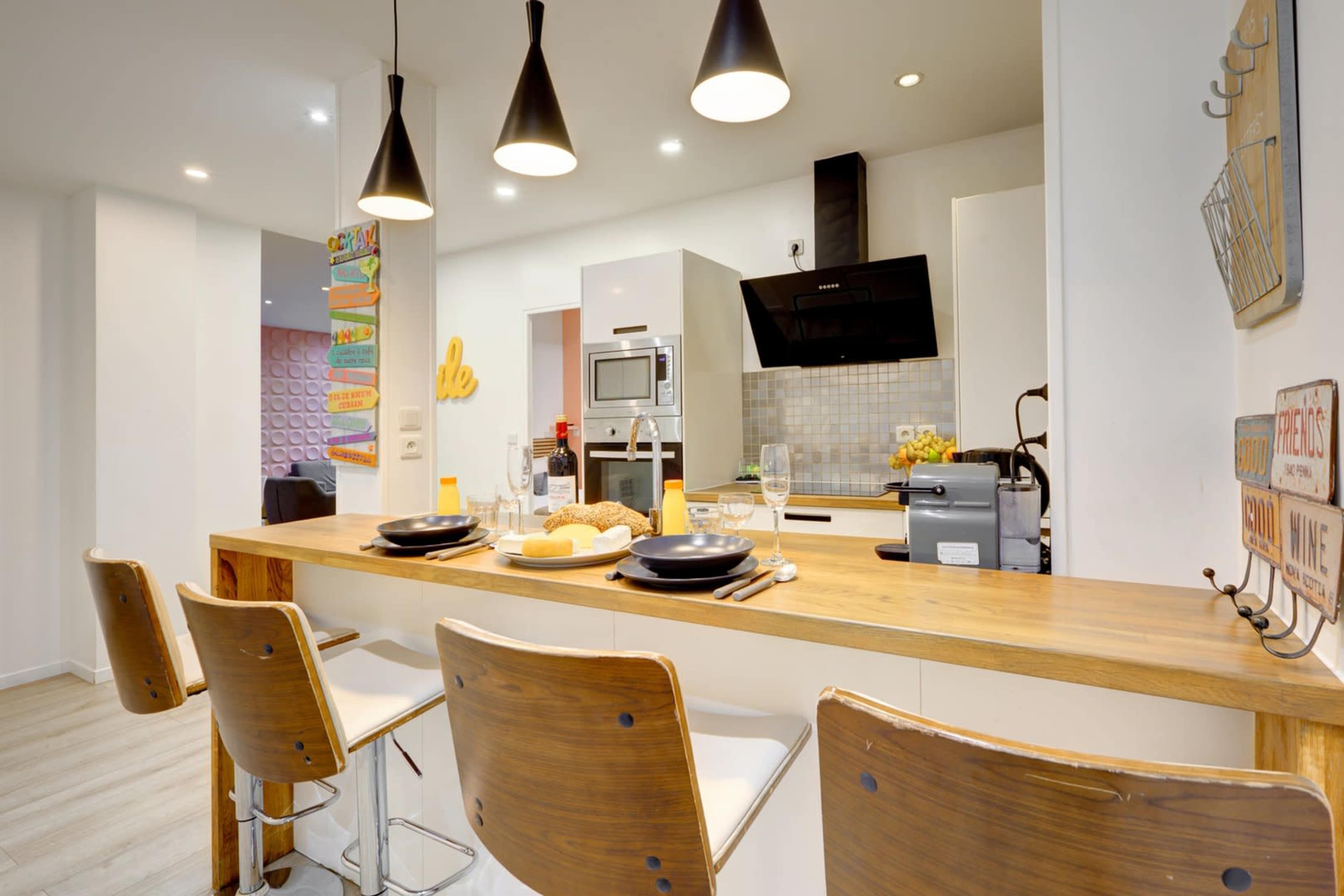 The image shows a modern kitchen with a wooden bar counter and three stools, featuring appliances such as a microwave and stovetop.