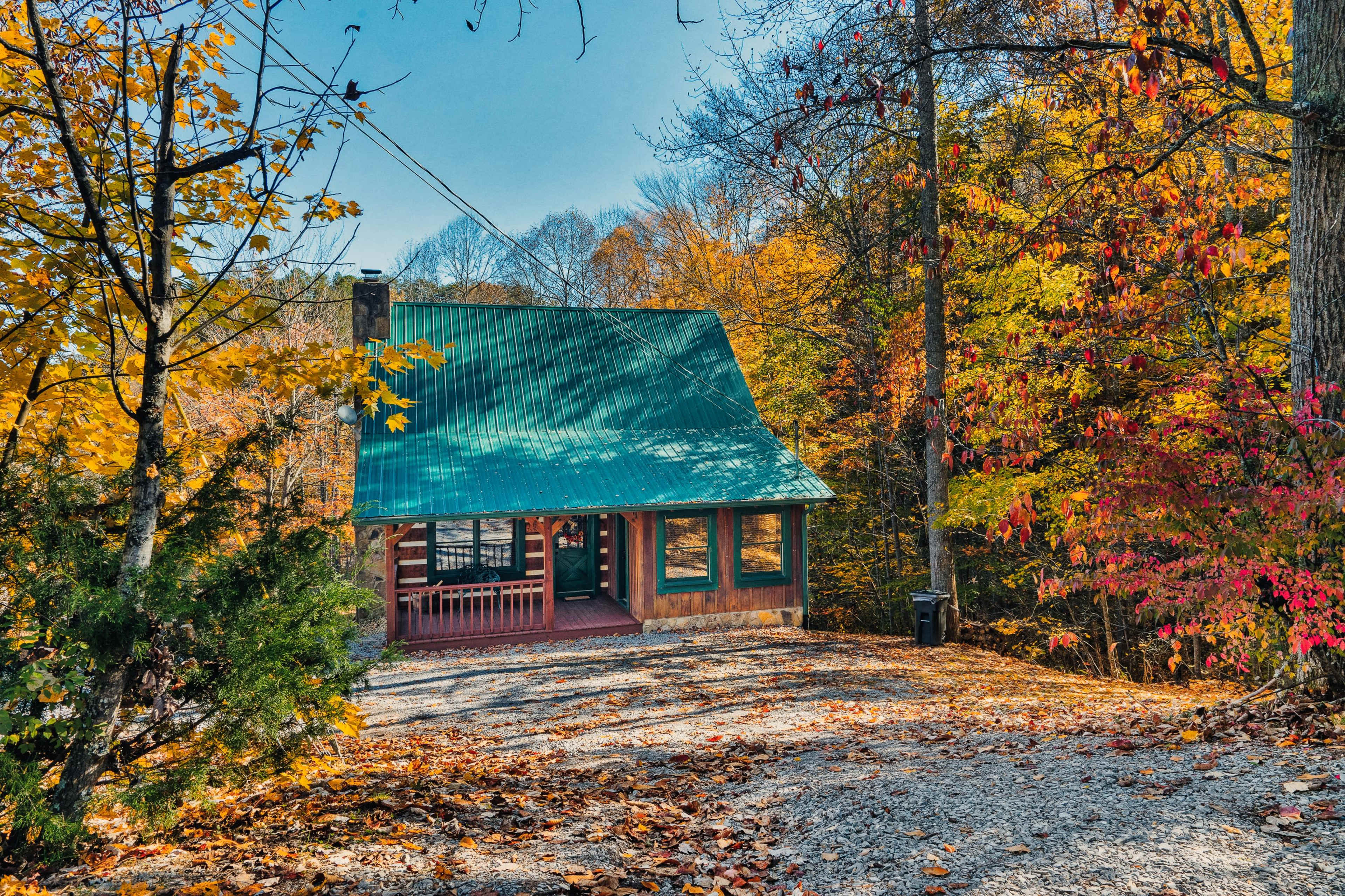 A cabin with a green metal roof sits on a gravel driveway surrounded by trees showcasing fall foliage.