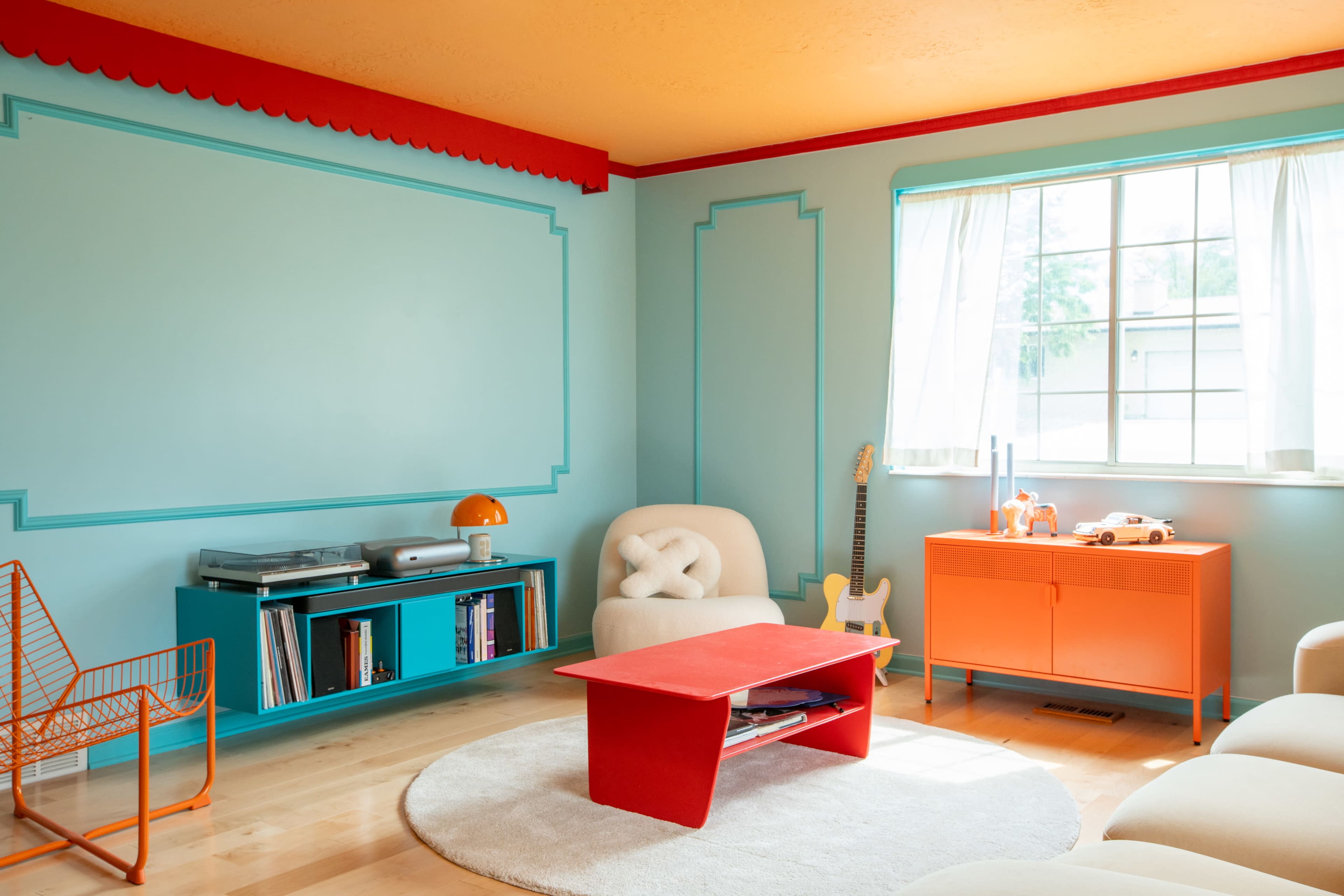 The room features a light blue wall with decorative trim, a bright orange ceiling, and colorful furniture including a red coffee table, orange cabinet, and a white chair.