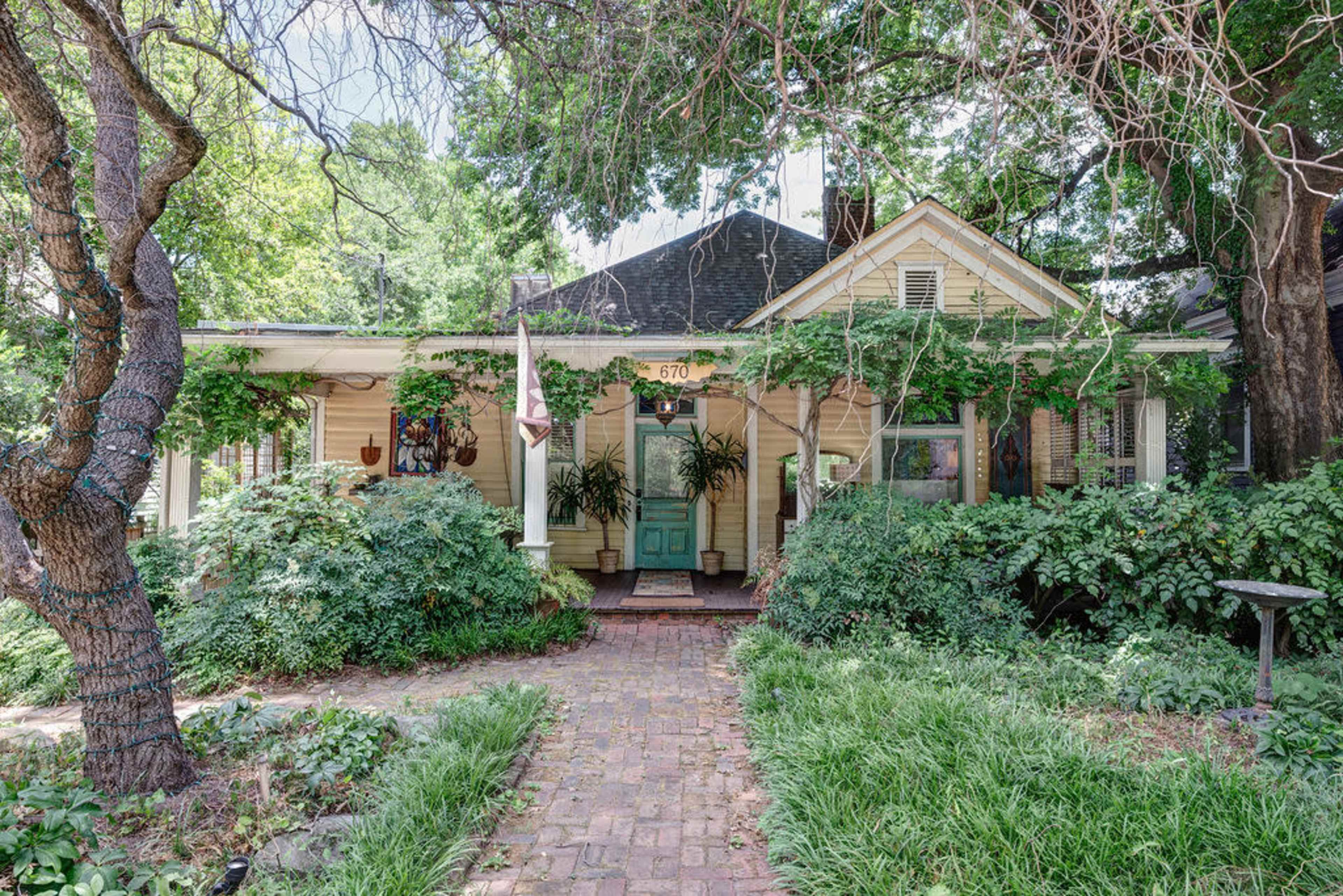 A charming yellow house features a covered porch with a teal front door, surrounded by lush greenery and flowering plants.