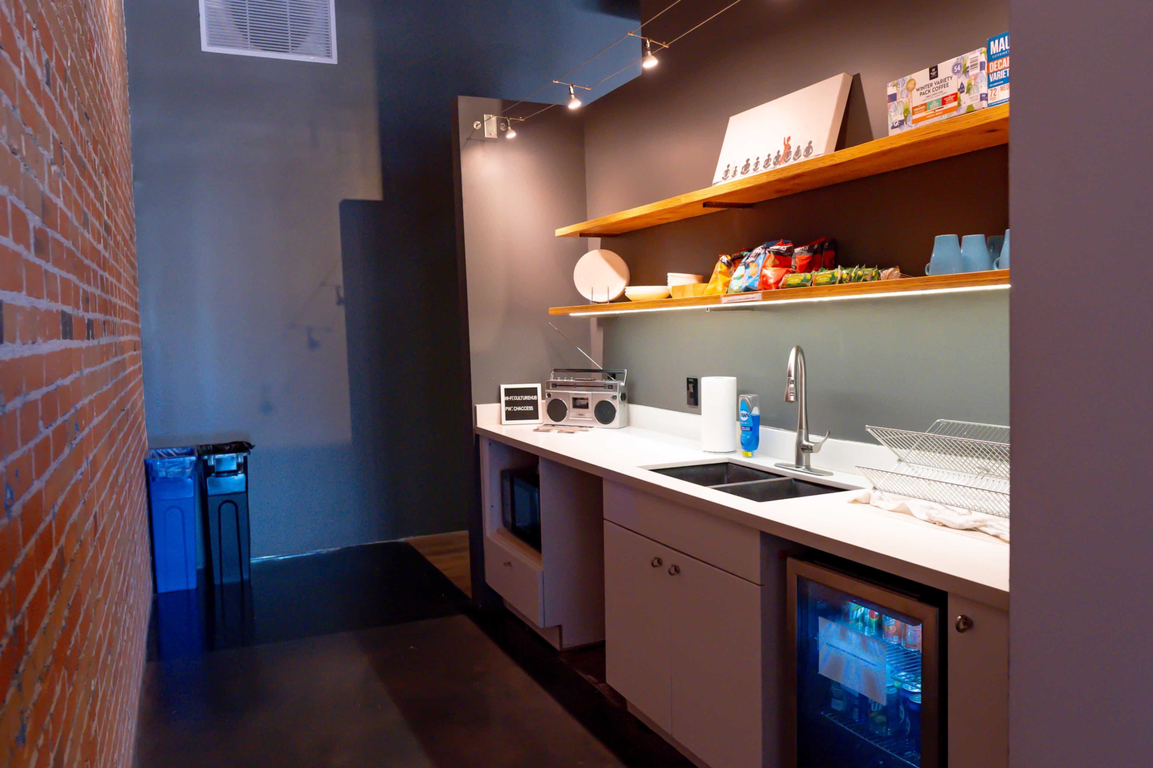 A modern kitchenette with countertops, a sink, a refrigerator, and shelves displaying various kitchen items, set against a brick wall.