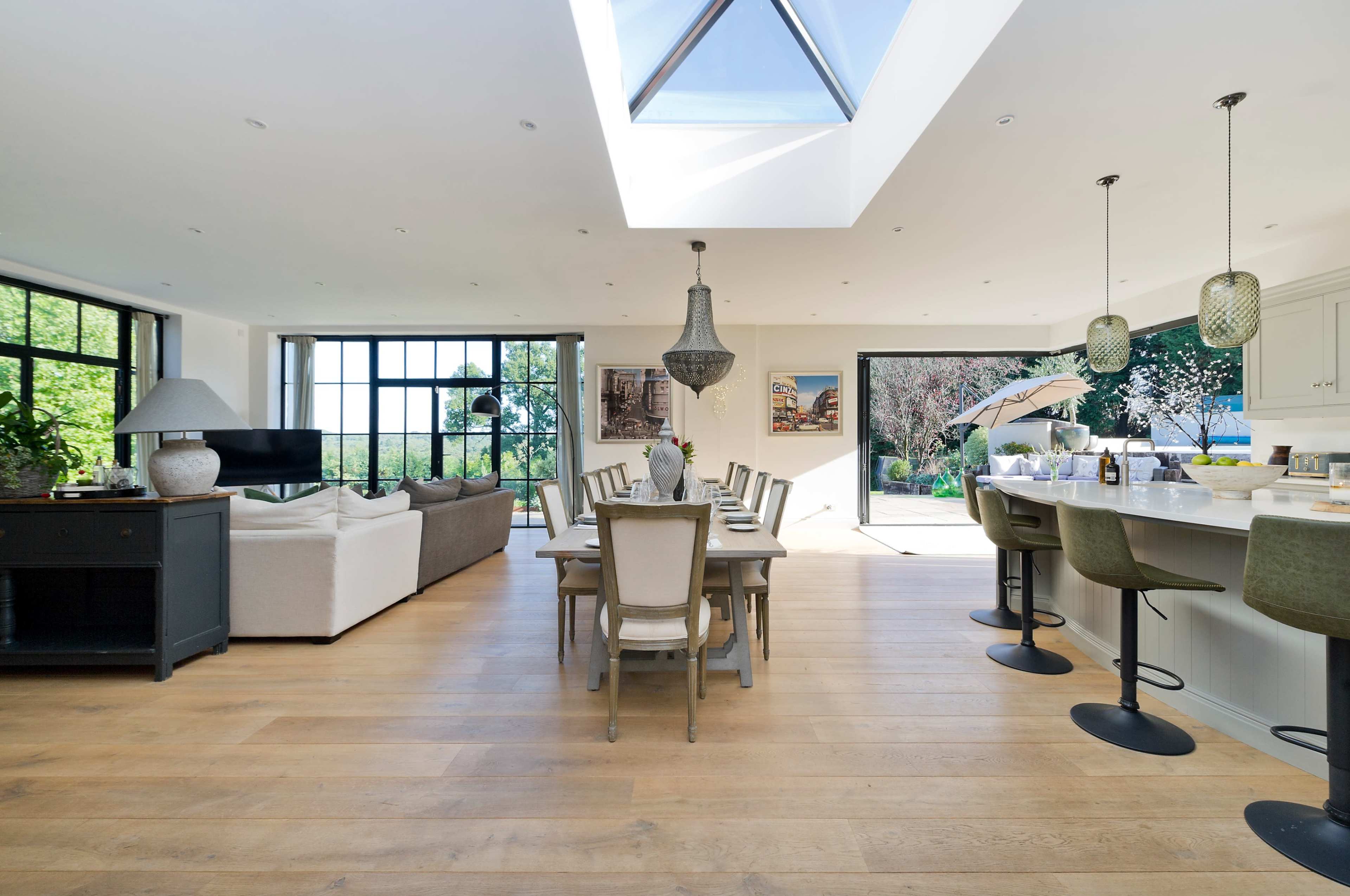 A spacious, modern open-plan living and dining area with a large skylight, a dining table set for eight, and a stylish kitchen bar with stools.