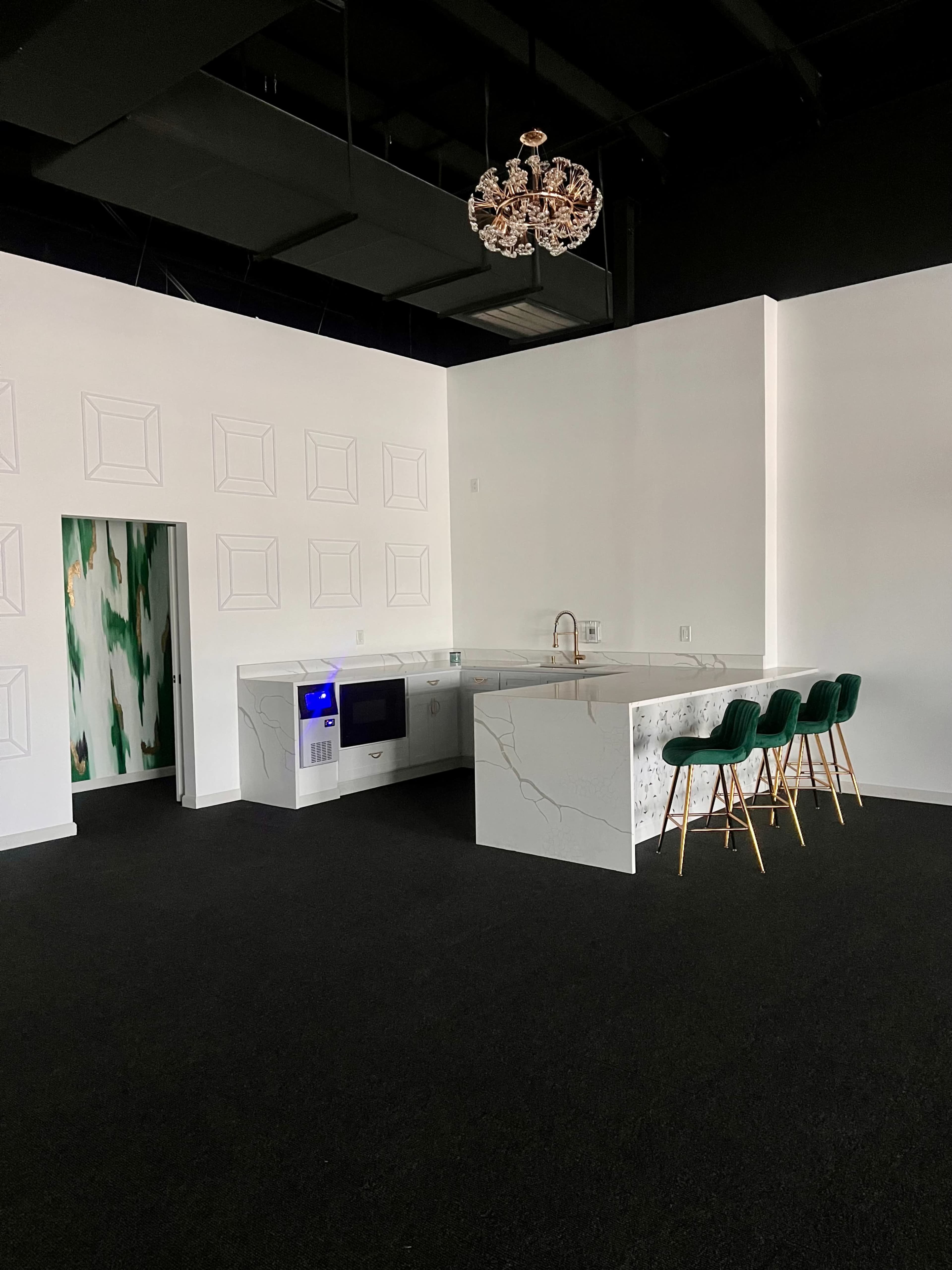 The image shows a modern kitchen with a white marble island, a microwave, and green velvet bar stools, set in a minimally decorated space with dark flooring and a decorative chandelier.