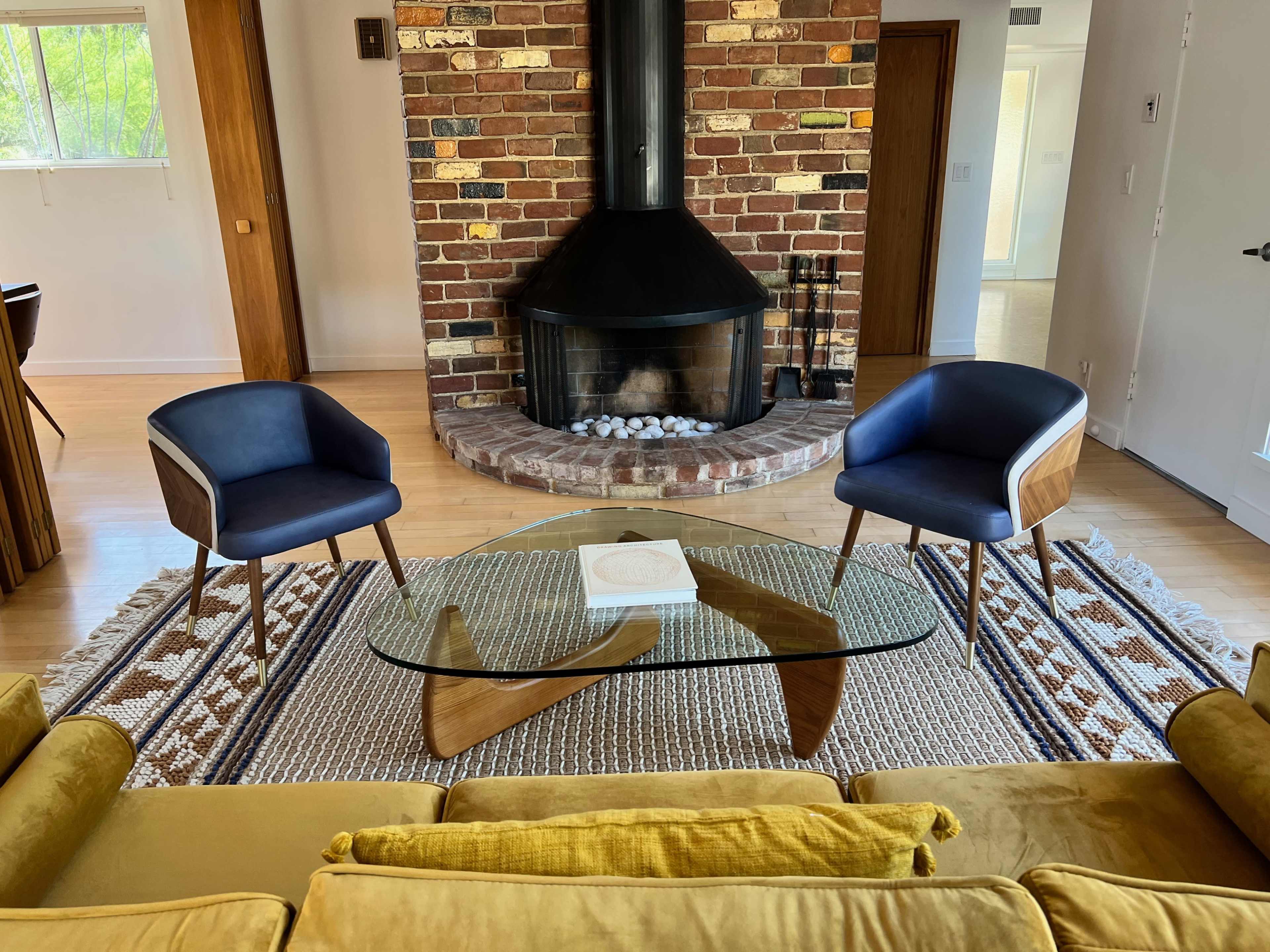 A modern living room features two blue chairs facing a glass coffee table in front of a brick fireplace.