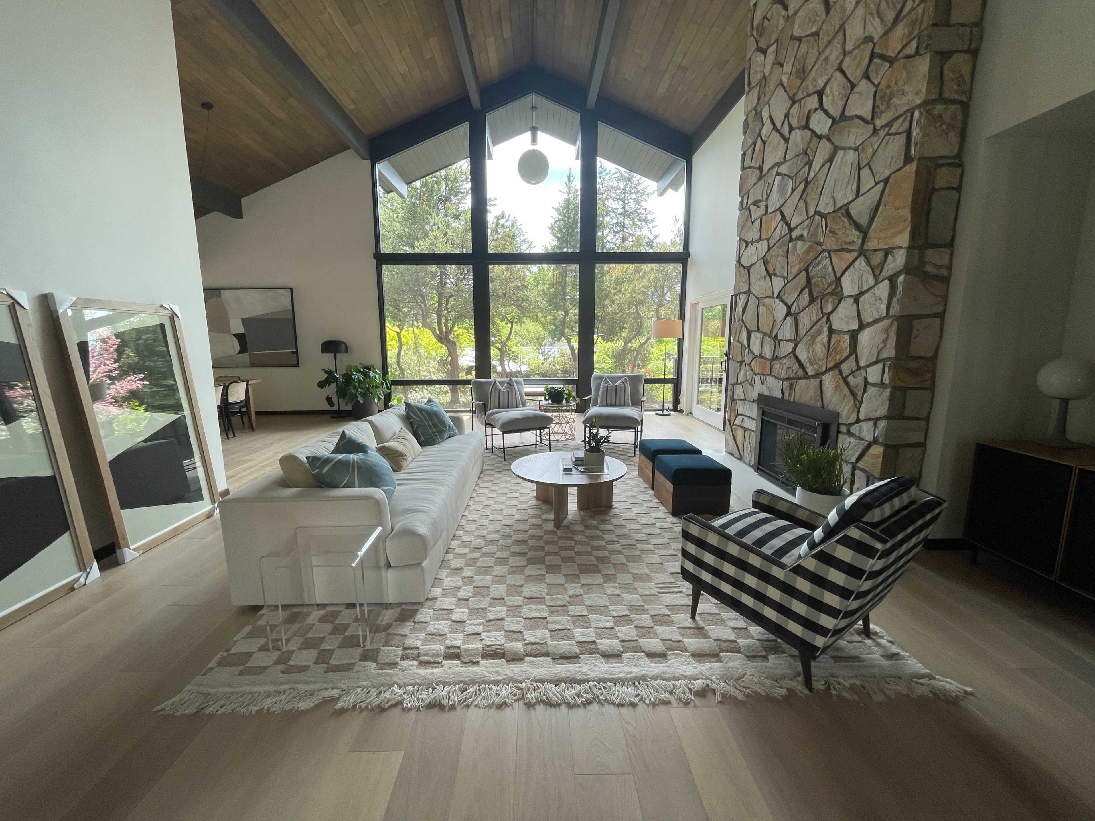 The scene features a spacious living room with a large stone fireplace, a mix of modern and patterned furniture, and large windows overlooking a garden.