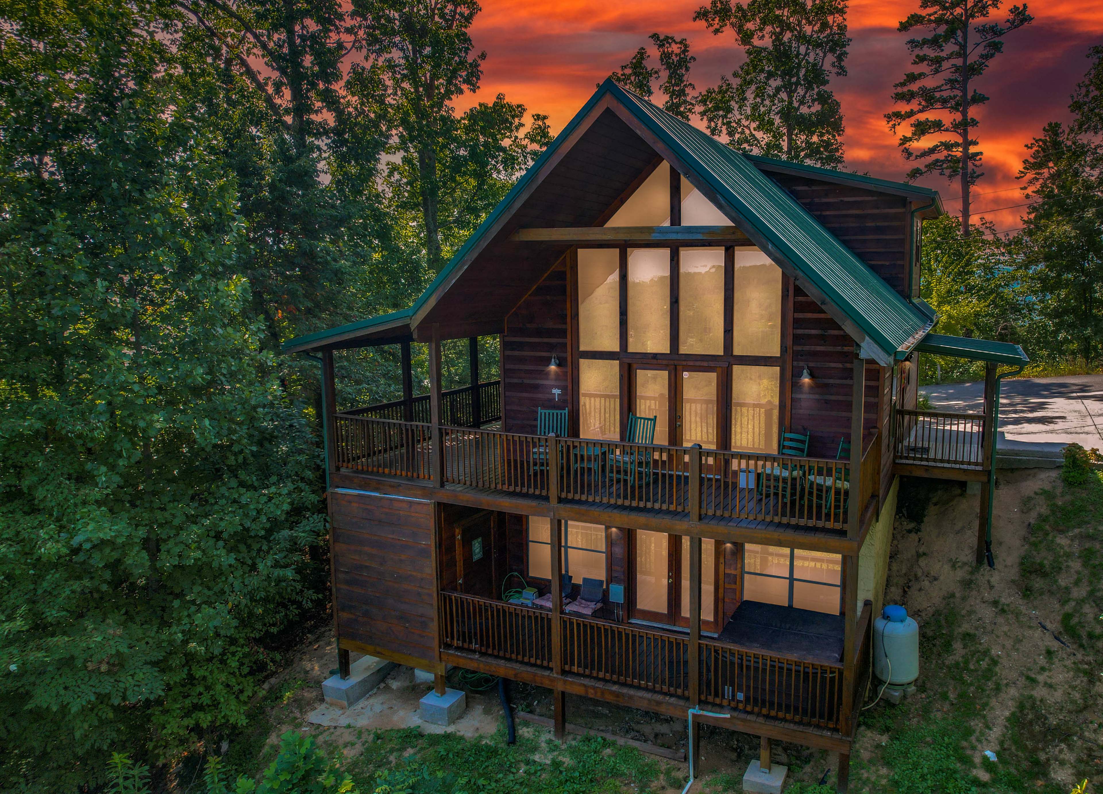 A two-story cabin with large glass windows and a green roof is situated among trees, overlooking a sloped hillside at sunset.