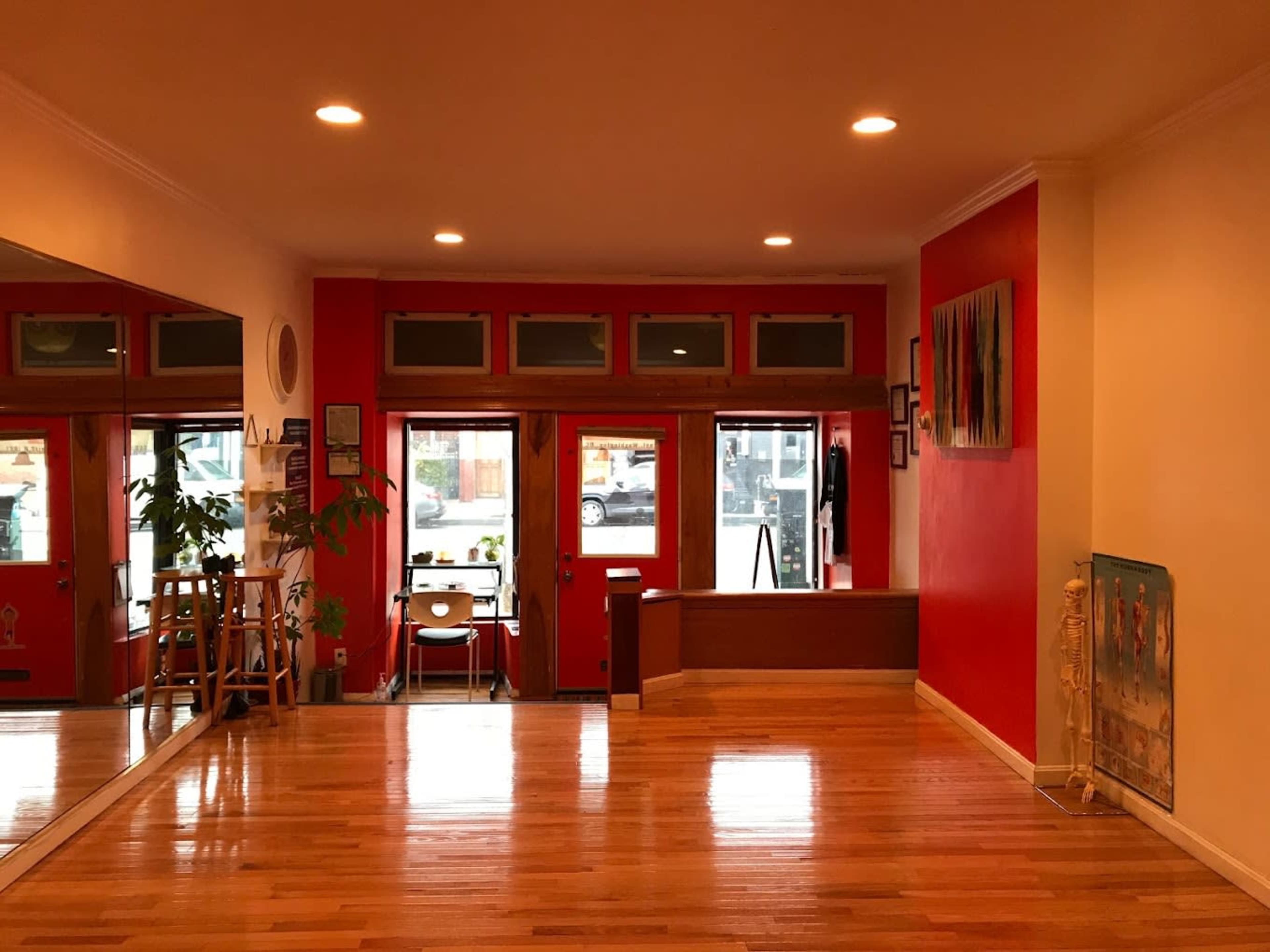 The image shows a spacious room with wooden floors, red walls, and large windows that allow natural light to enter.